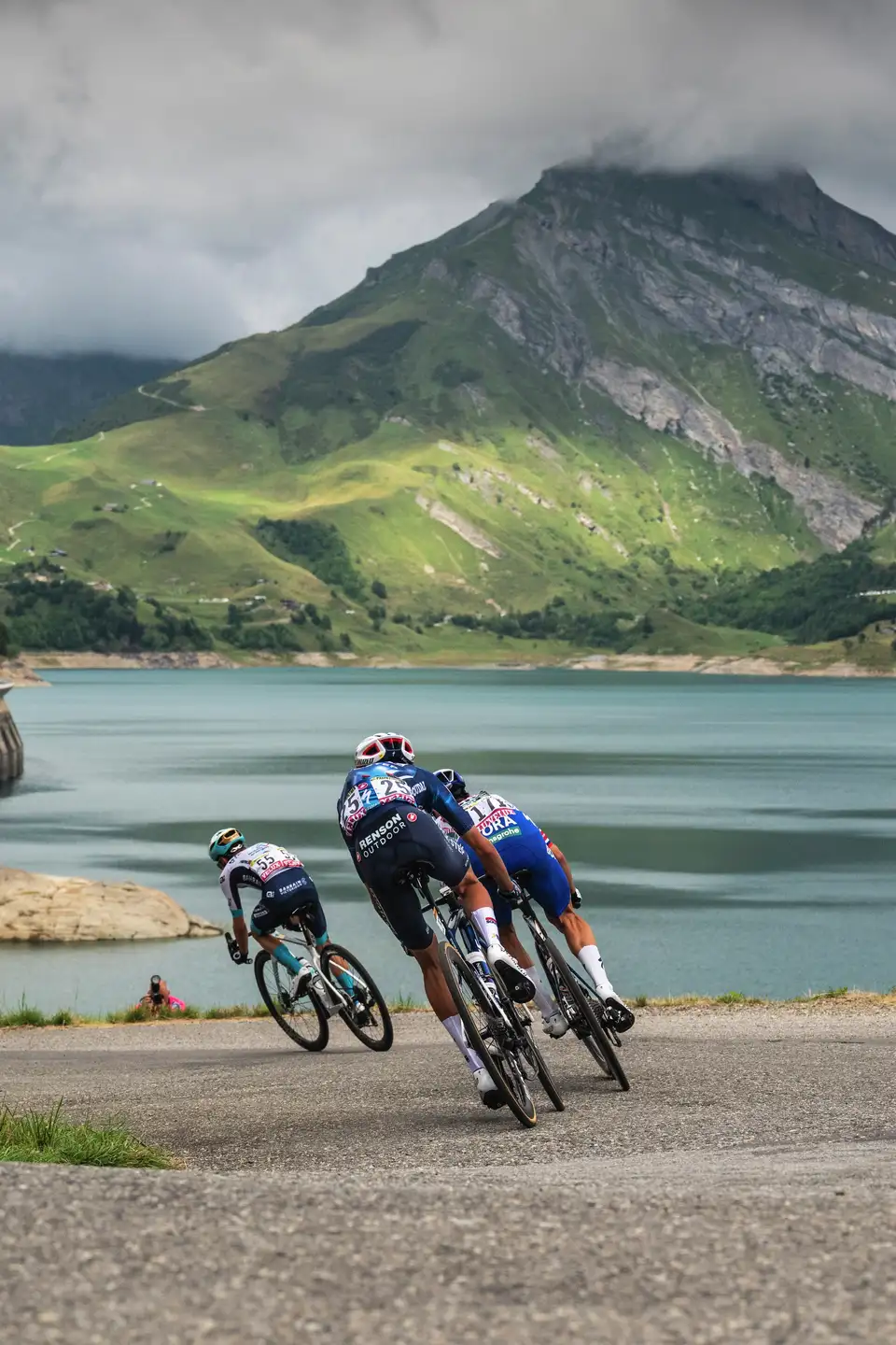 Tour de France-ryttere, der kører langs en bjergvej ved en sø under en overskyet himmel.