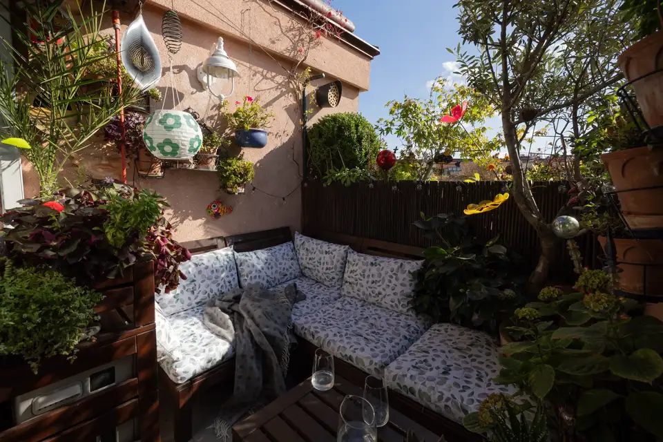 Angolo salotto esterno confortevole in un giardino, con una panchina in legno, cuscini decorati, piante verdi e fiori. Decorazioni sospese e fioriere aggiungono fascino, creando un’atmosfera accogliente per il relax o le riunioni.
