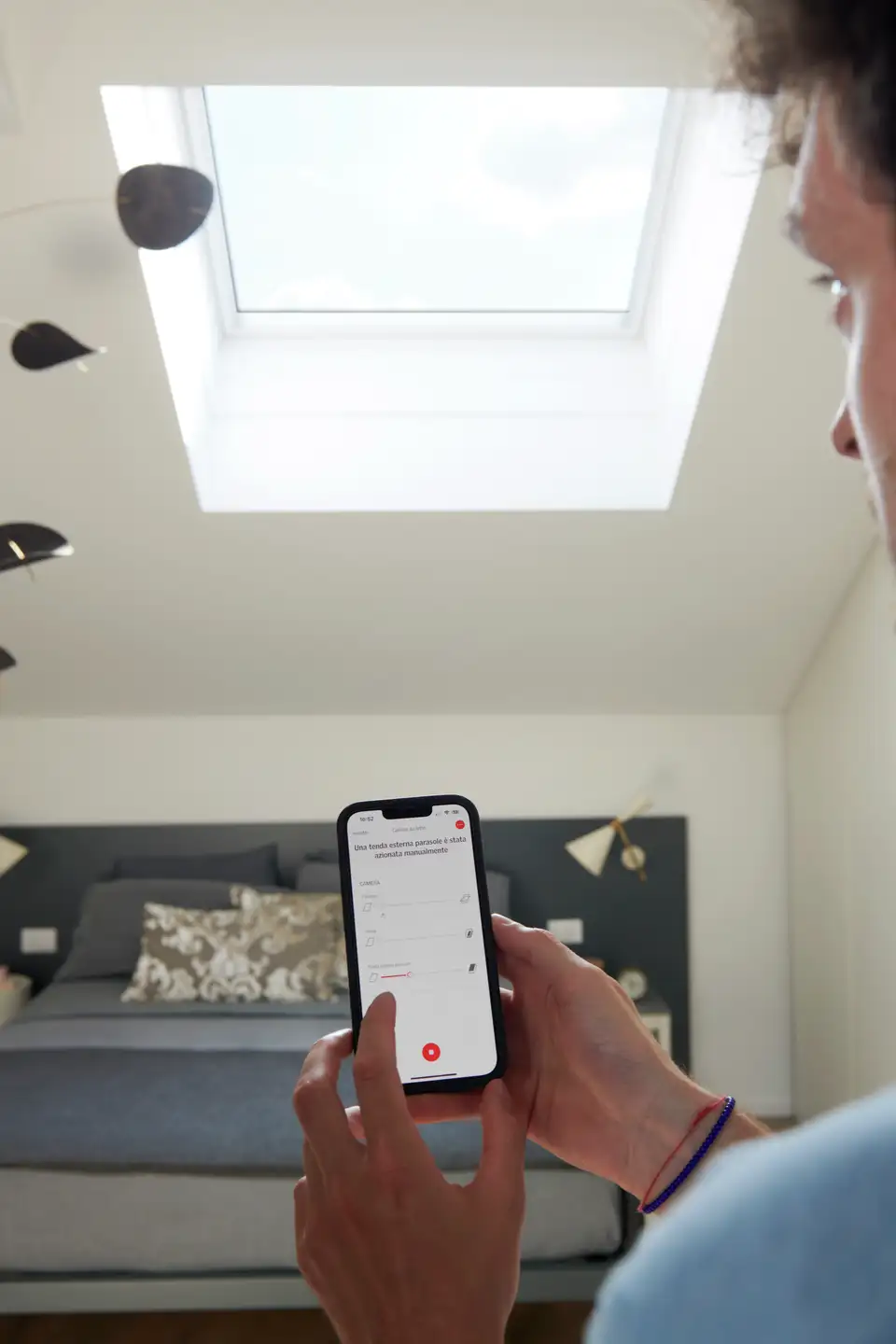 Osoba ovládající střešní okno pomocí aplikace na smartphonu v moderní ložnici. | VELUX