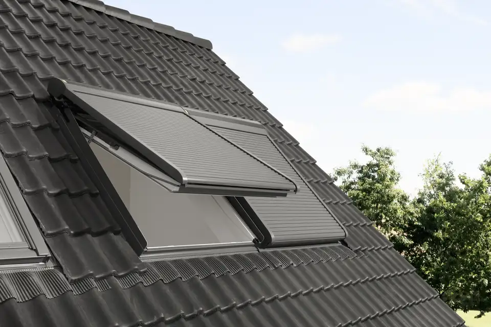 Toit avec fenêtre de toit et volet roulant VELUX partiellement ouvert, tuiles foncées et arbres visibles