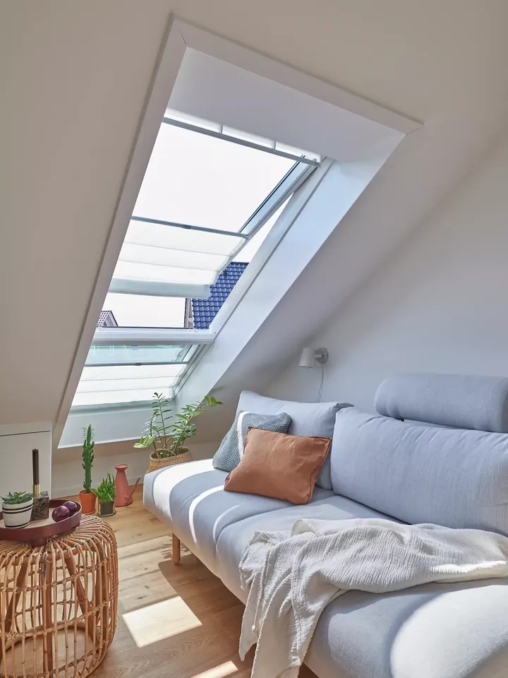 Wohnzimmer im Dachboden mit grauem Sofa, Pflanzen und VELUX Dachflächenfenster, durch das Sonnenlicht einfällt.