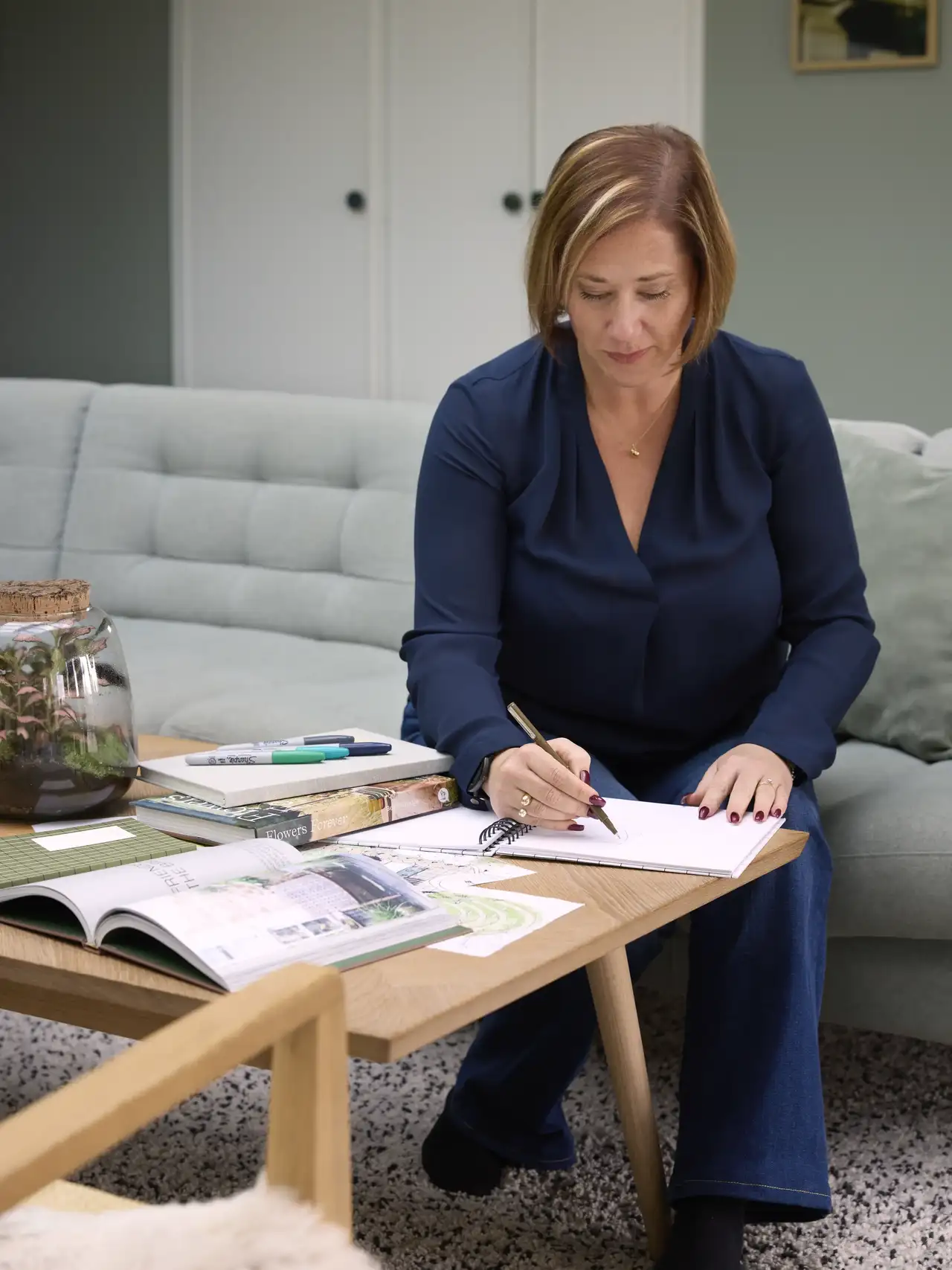 Kvinne som sitter og skriver notater ved sofabordet i en moderne innredet stue. På bordet ligger bøker og tidsskrifter i tillegg til at det står en plante i en glasskuppel.