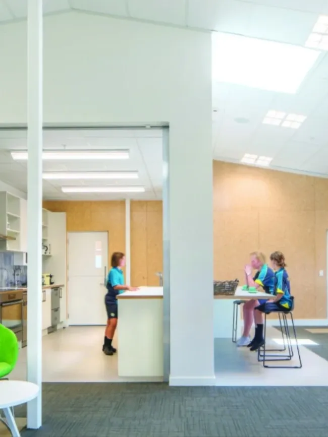 Open school common room with kitchen, students at counter, and bright green lounge chairs.