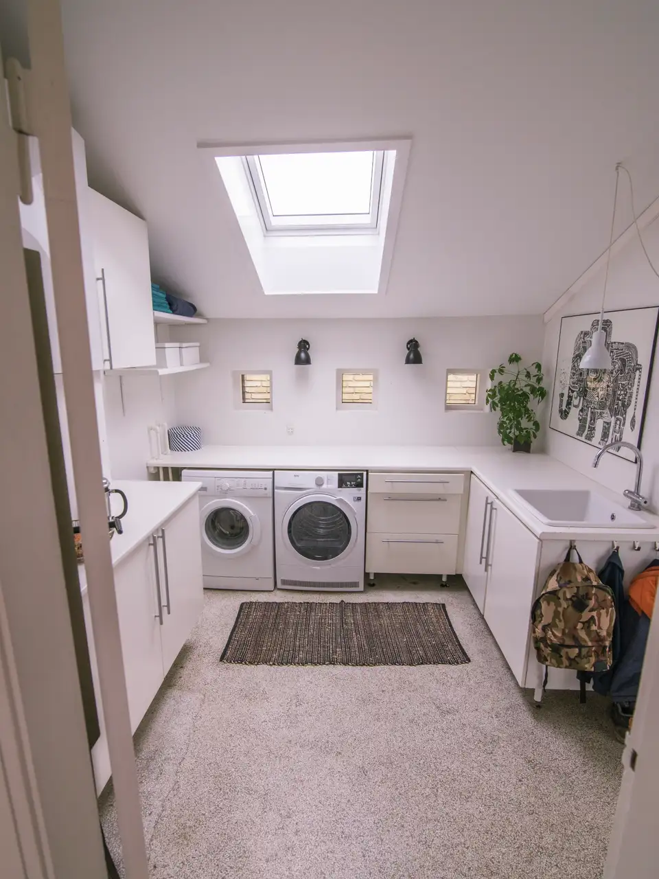 buanderie mansardée avec fenêtre de toit VELUX au-dessus du lave-linge et du sèche-linge, meubles de rangement et vasque répartis le long des murs