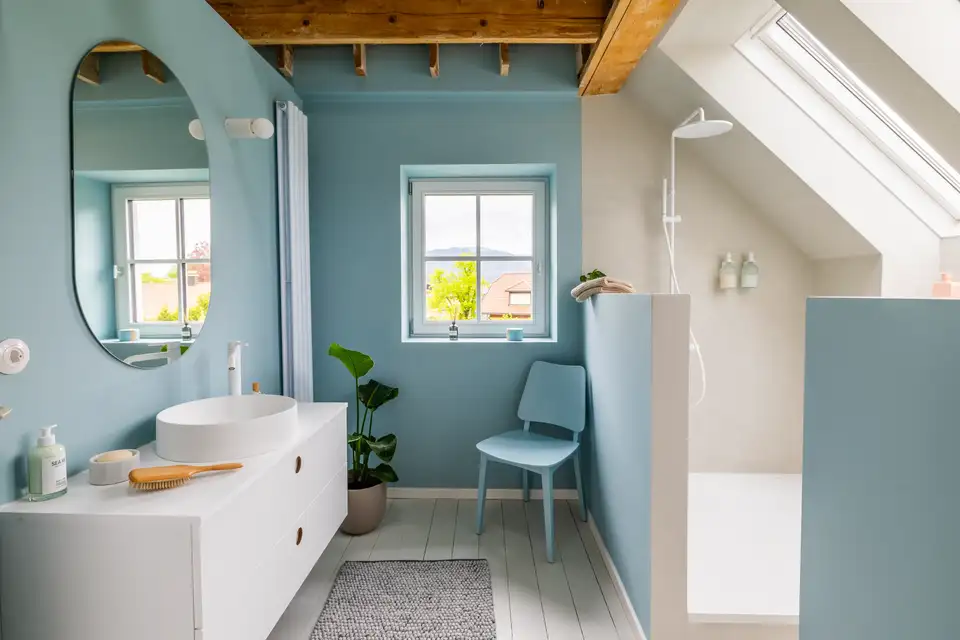 Salle de bains lumineuse avec murs bleu clair, siège assorti et fenêtres de toit VELUX diffusant une lumière douce et harmonieuse. 