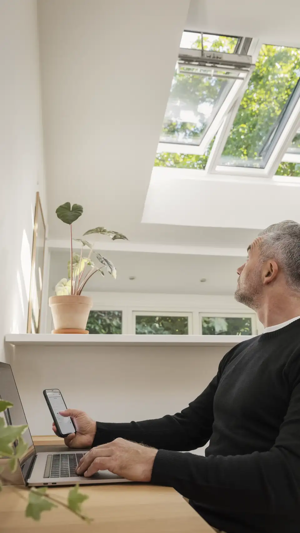 Una persona nel suo spazio di lavoro casalingo controlla le finestre per tetti VELUX dal suo smartphone con l’App VELUX