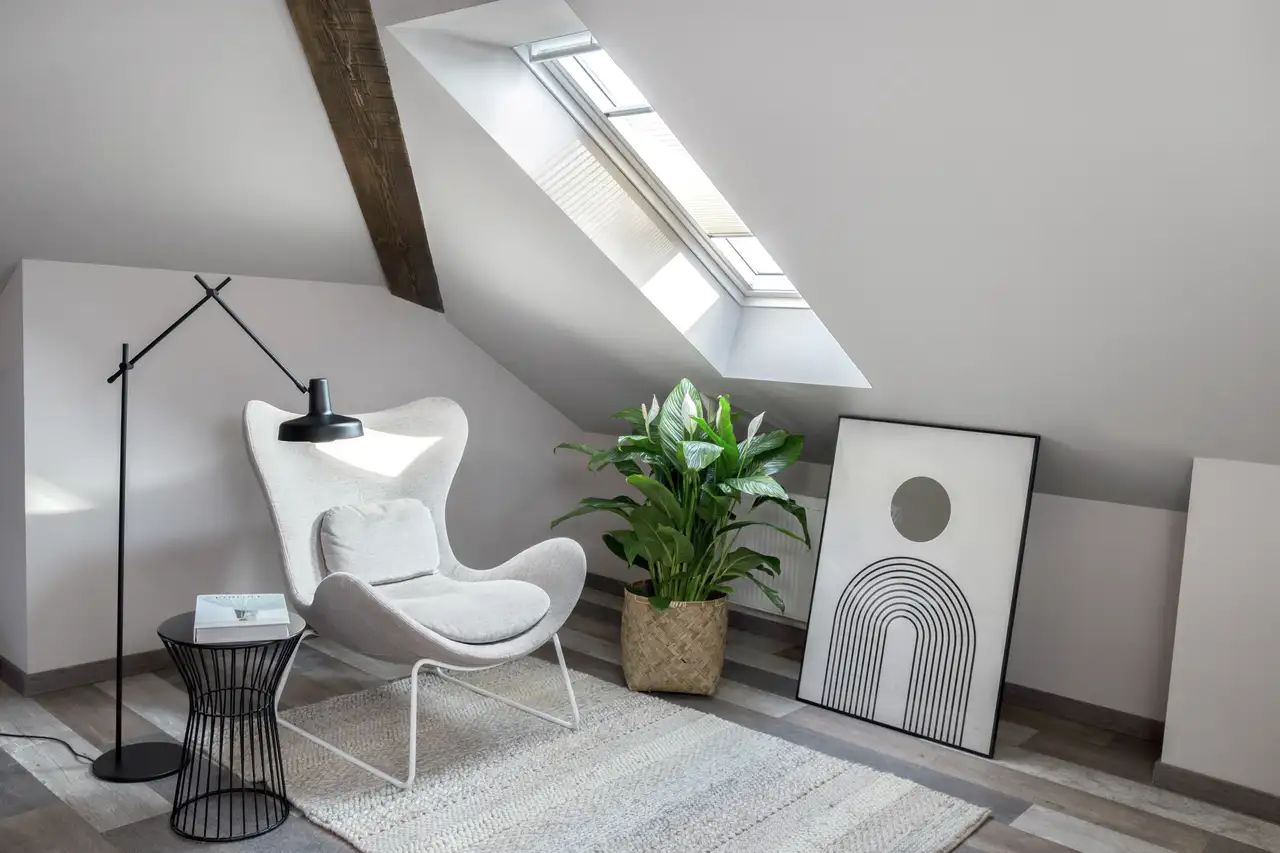 Fauteuil blanc dans un coin détente sous une fenêtre de toit, accompagné d’une lampe, d’une plante et d’un tableau.