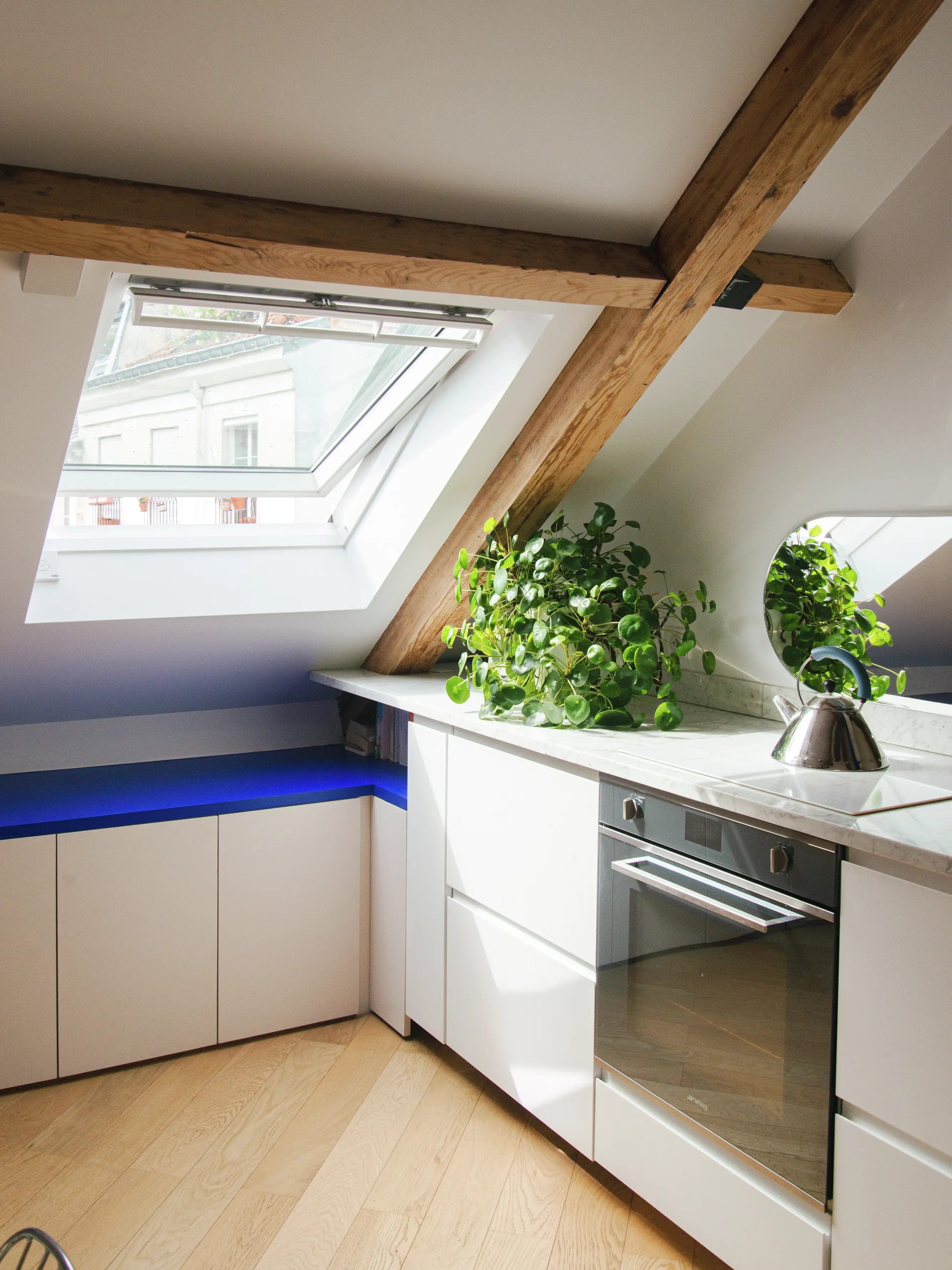 Cuisine moderne sous les combles avec des poutres en bois apparentes, des meubles blancs, un éclairage d'ambiance bleu et une fenêtre de toit apportant de la lumière naturelle.