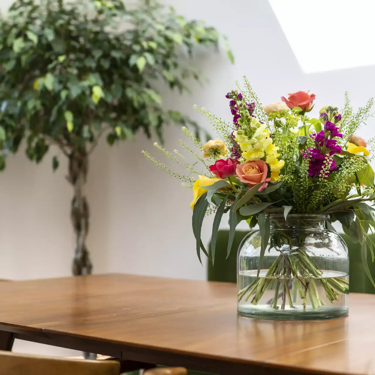 Frisches Blumenarrangement auf hölzernem Esstisch mit unscharfem Pflanzenhintergrund.
