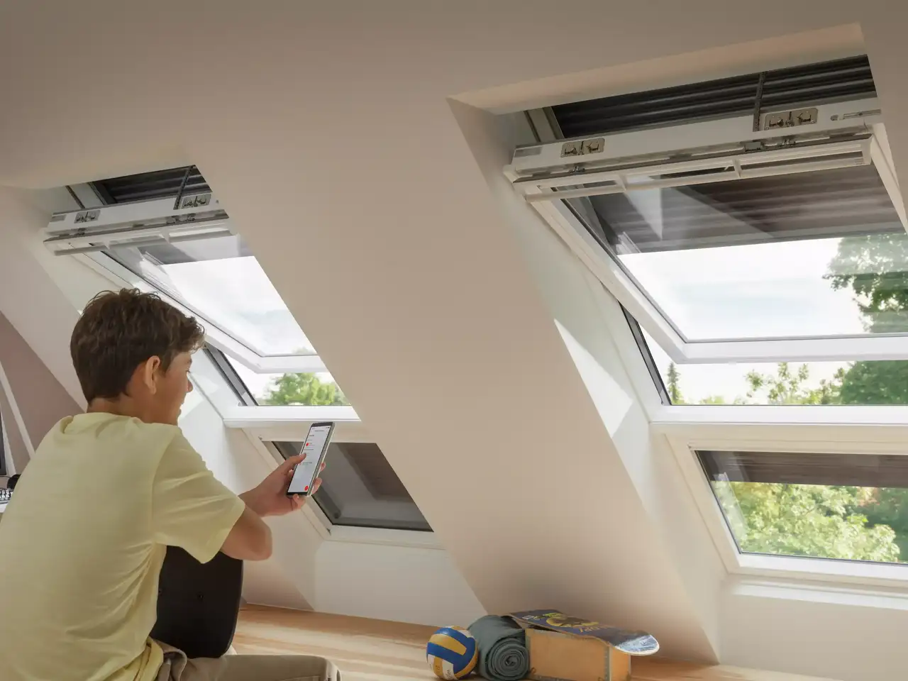 Person verwendet Smartphone zur Kontrolle eines VELUX Dachflächenfensters in einem hellen Dachbodenraum.