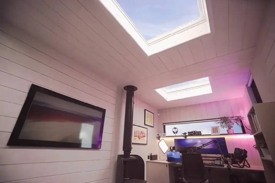 Intérieur avec plafond incliné en bois et deux puits de lumière. À gauche, une télévision murale affichant une image abstraite. En dessous, un poêle à bois noir. À droite, un bureau avec plusieurs moniteurs et des haut-parleurs. Éclairage ambiant rose-violet, créant une atmosphère moderne et cosy.
