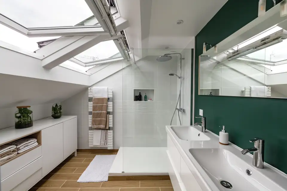Salle de bain mansardée équipée de fenêtres de toit motorisées VELUX ouvertes, offrant lumière naturelle et ventilation optimale.
