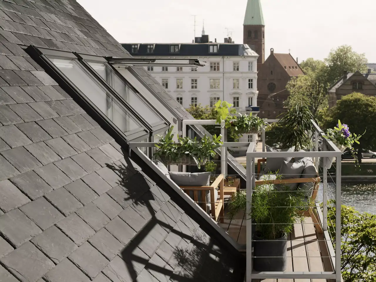 L’image montre une terrasse sur le toit avec un plancher en bois et une balustrade en verre, offrant un espace extérieur au sommet d’un bâtiment. Il y a des plantes et des meubles d’extérieur, suggérant que cet espace est utilisé pour les loisirs. En arrière-plan, on voit d’autres bâtiments et des arbres verts, indiquant un environnement urbain.