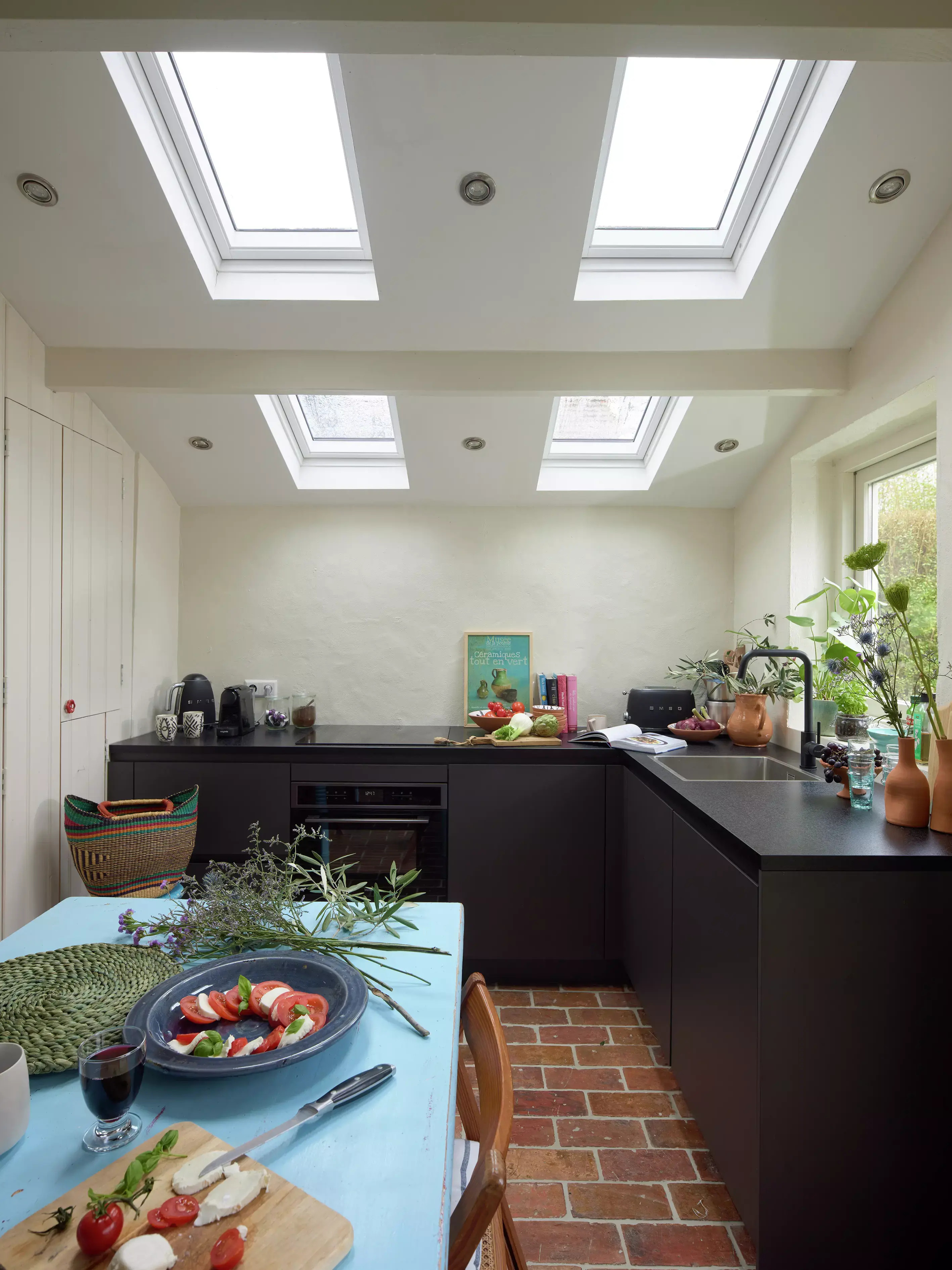 Cuisine moderne avec des fenêtres de toit VELUX, des comptoirs noirs, un sol en brique et une installation de repas frais.