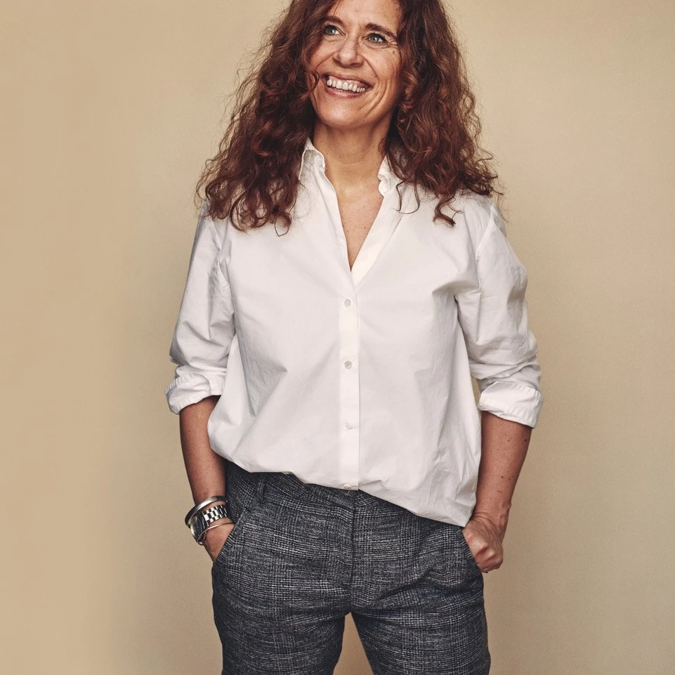 Donna sorridente con camicia bianca e pantaloni grigi, le mani nelle tasche, davanti a uno sfondo beige | Rivista VELUX