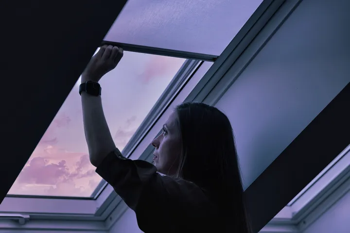 Žena zatvara roletu na krovnom prozoru u sumrak, s ljubičastim nebom u pozadini | VELUX