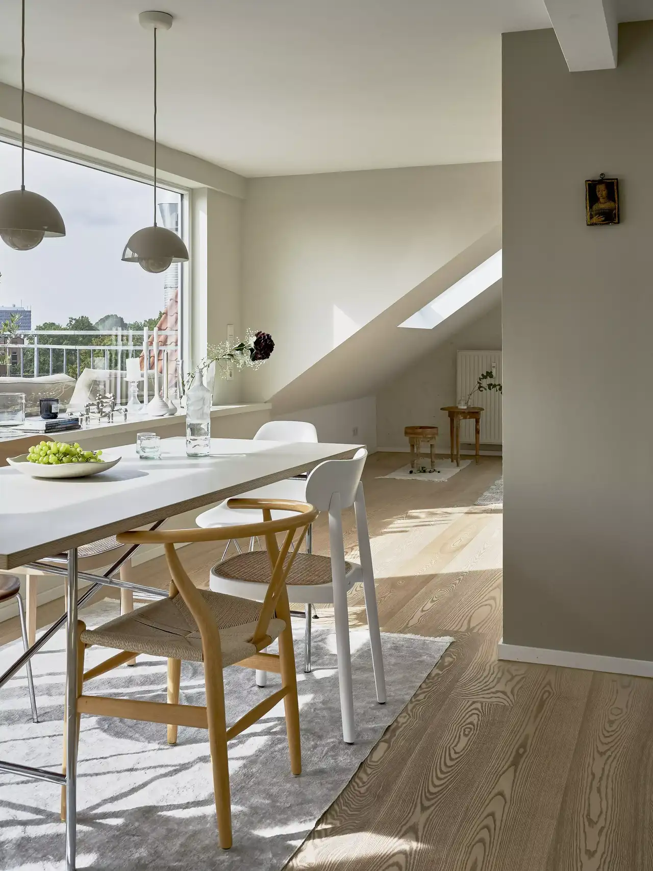 Une salle à manger moderne avec une table blanche et des chaises en bois. Des suspensions sont accrochées au-dessus de la table. La pièce est lumineuse grâce aux grandes fenêtres. Un cadre photo est accroché au mur et un tapis gris est sous la table.