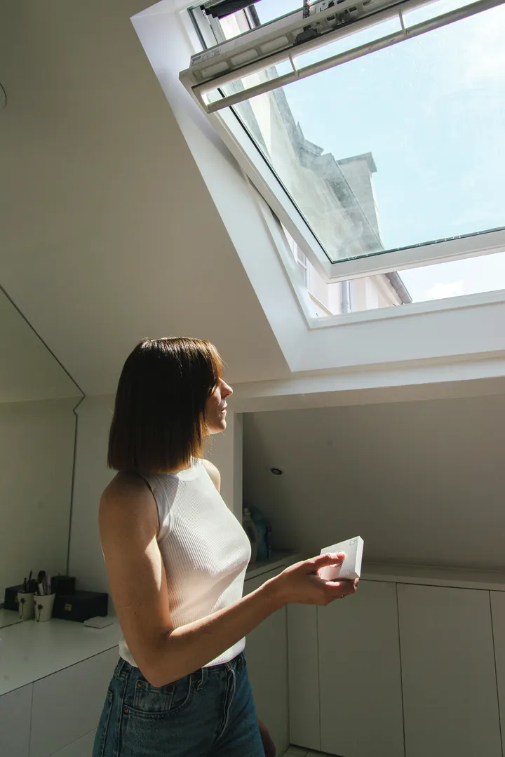 Žena koristi daljinski upravljač za otvaranje krovnog prozora, puštajući svež vazduh i sunčevu svetlost u moderno potkrovlje kupatilo. | VELUX 