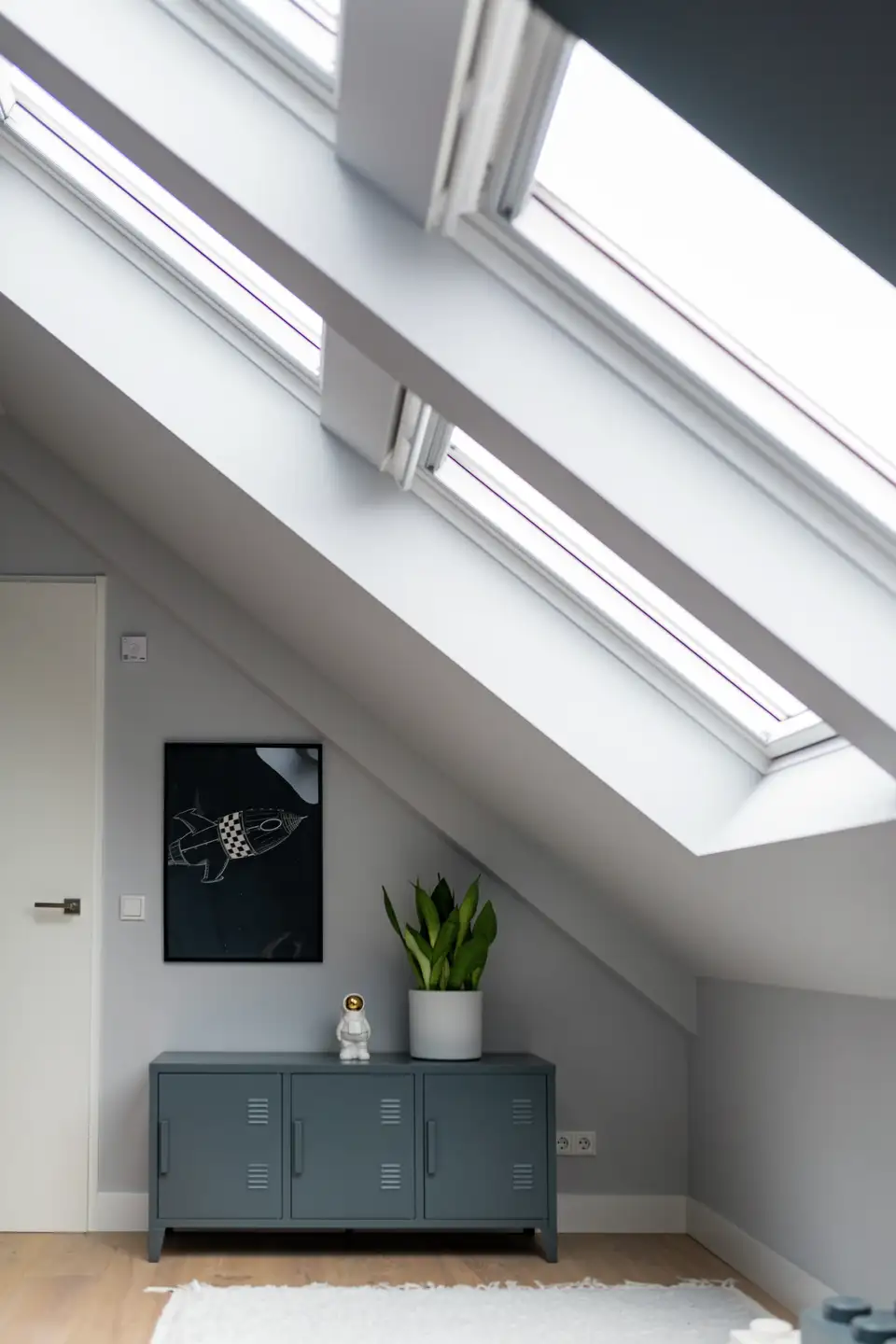Minimalistički potkrovni prostor s više krovnih prozora, prirodnim svjetlom koje ispunjava sobu i modernim dekorom koji uključuje sivi ormar i biljku. | VELUX