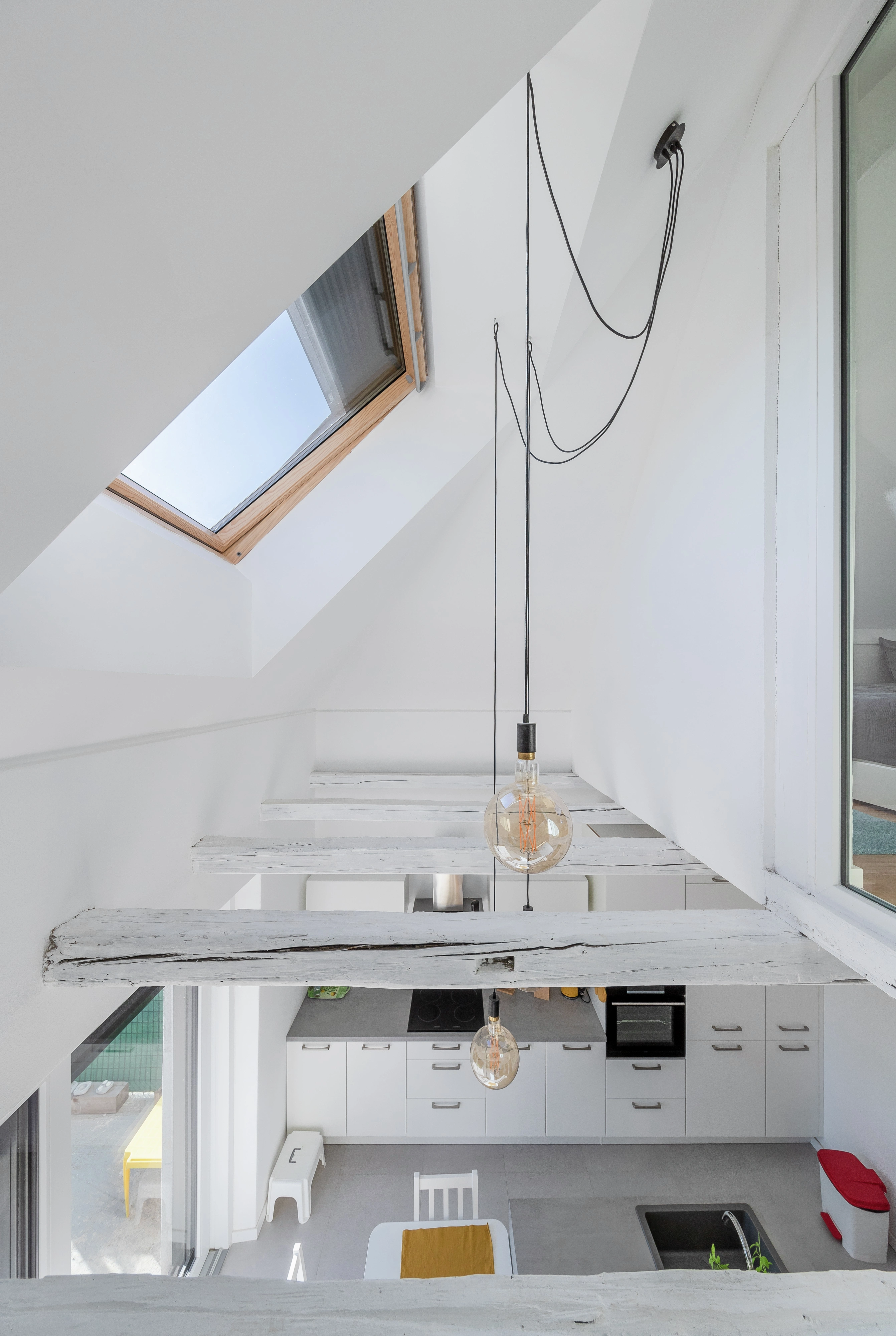 Nyitott tetőtéri tér látszó fa gerendákkal, függesztett világítással és VELUX tetőablakkal, amely bőséges természetes fényt enged be a konyha fölötti térbe és az üvegfalú szülői hálóba