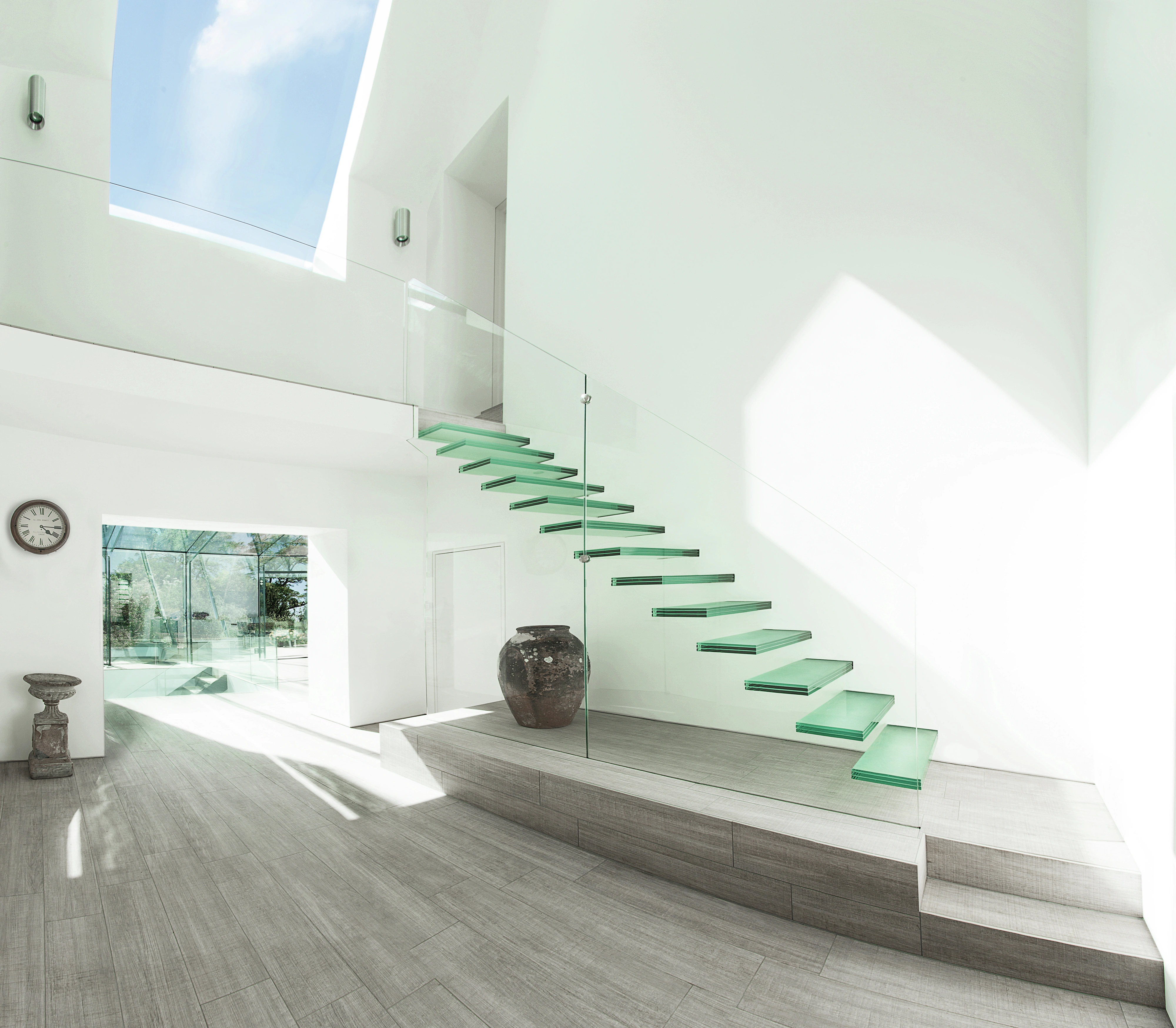 L’immagine mostra uno spazio interno con un design moderno caratterizzato da una scala con ringhiere in vetro e gradini con una tinta verde. C’è un lucernario sopra che permette alla luce naturale di riempire la stanza. Lo spazio appare pulito e minimalista, il che potrebbe essere interessante per scopi architettonici o di design d’interni.