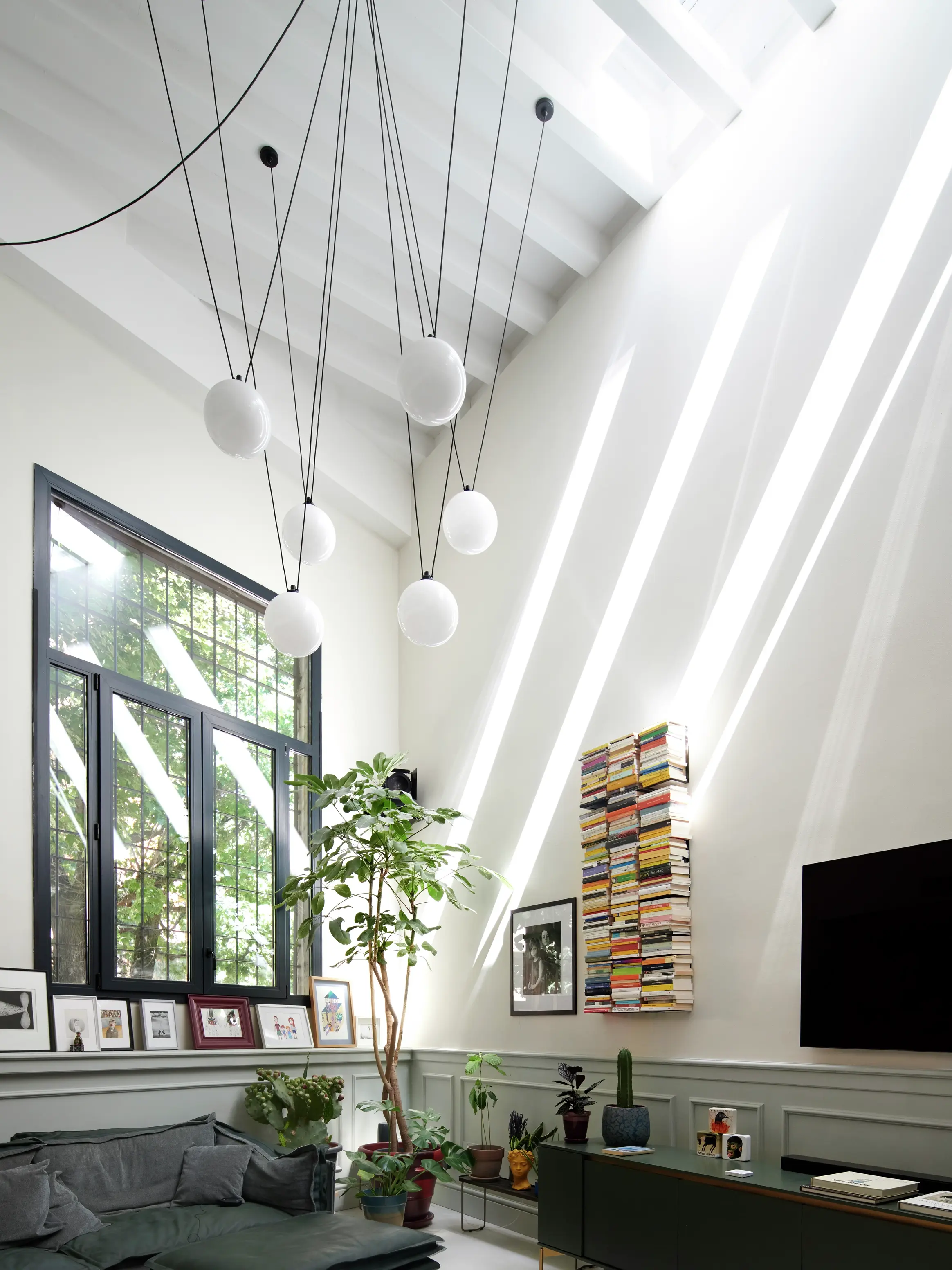 Salon moderne avec des fenêtres de toit VELUX, des suspensions, des plantes et une bibliothèque.