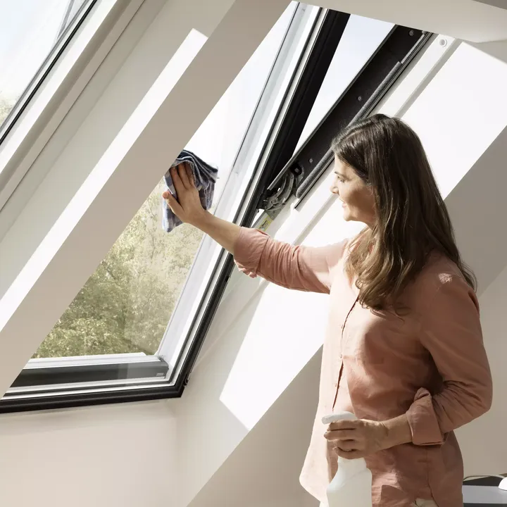 Person reinigt ein VELUX Dachflächenfenster mit einem Tuch, helles Tageslicht scheint hindurch.