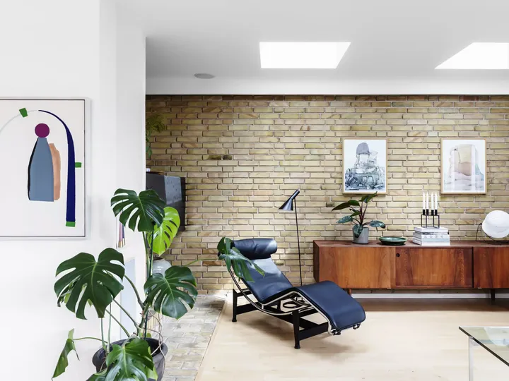 Stilvolles Wohnzimmer mit schwarzer Chaiselongue, hölzernem Sideboard, Ziegelwand und grünen Pflanzen.