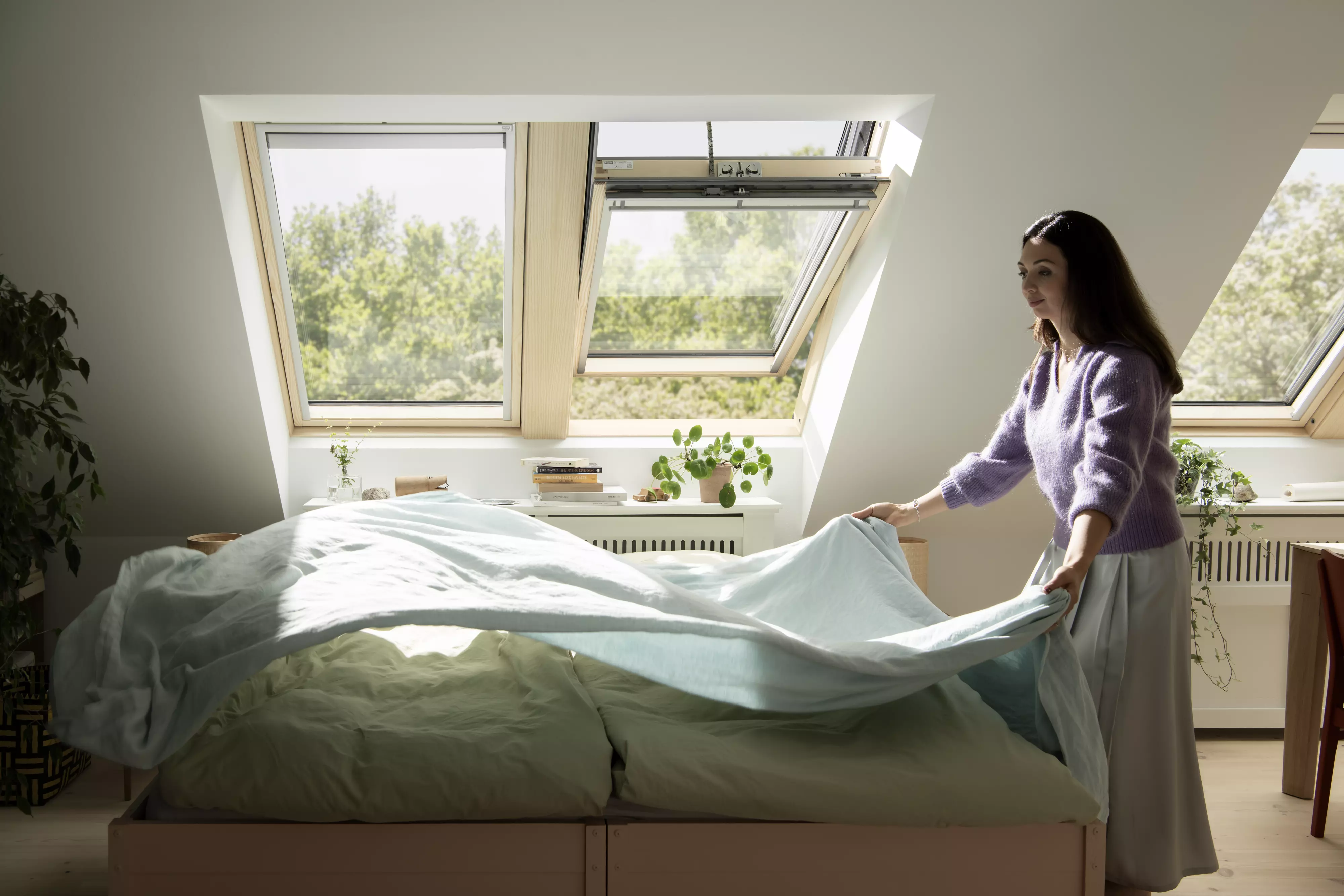 Bright bedroom with open VELUX roof windows and a person arranging the bed.