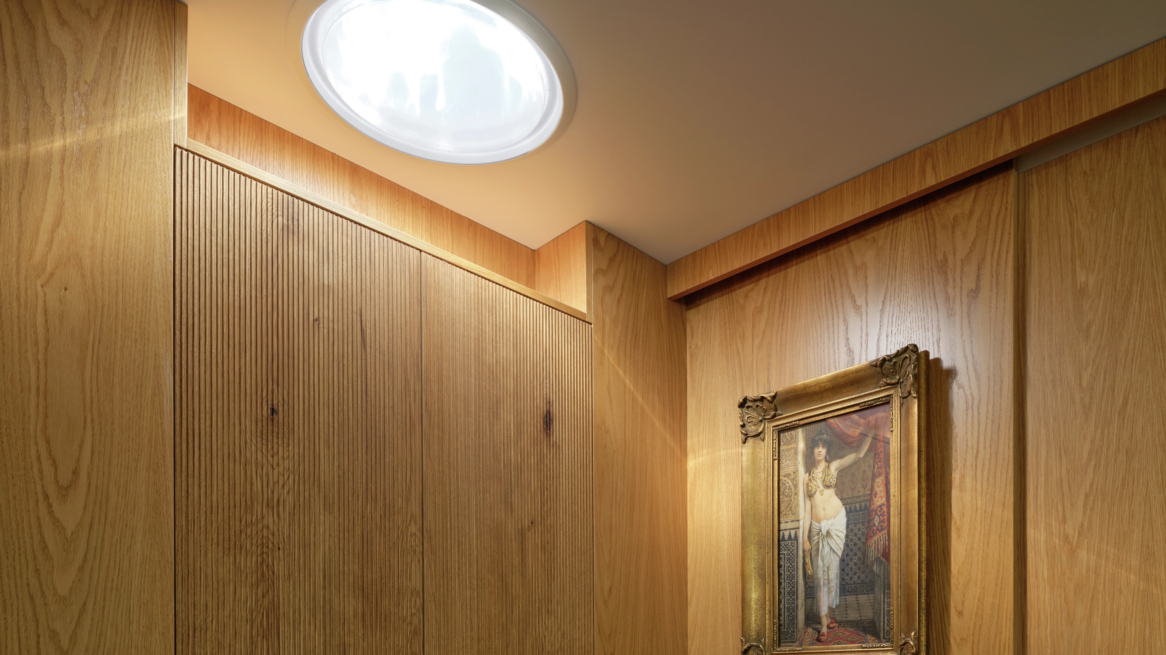 Wood-paneled hallway lit by a round sun tunnel above a framed painting.