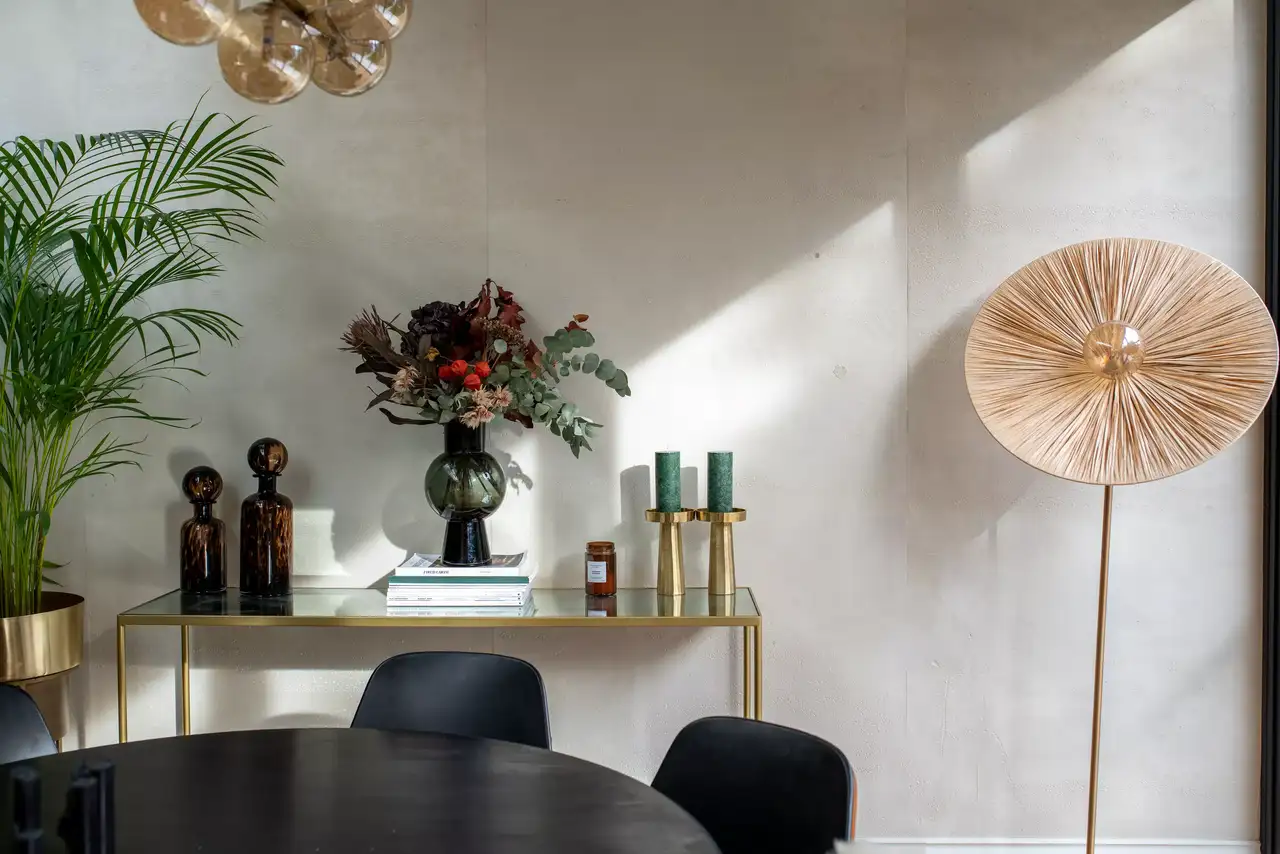 Intérieur élégant avec un mur clair, une table ronde noire au premier plan, et un buffet doré sur lequel repose un vase avec des fleurs, des bougies et des livres. À droite, un lampadaire circulaire crée une ambiance chaleureuse.