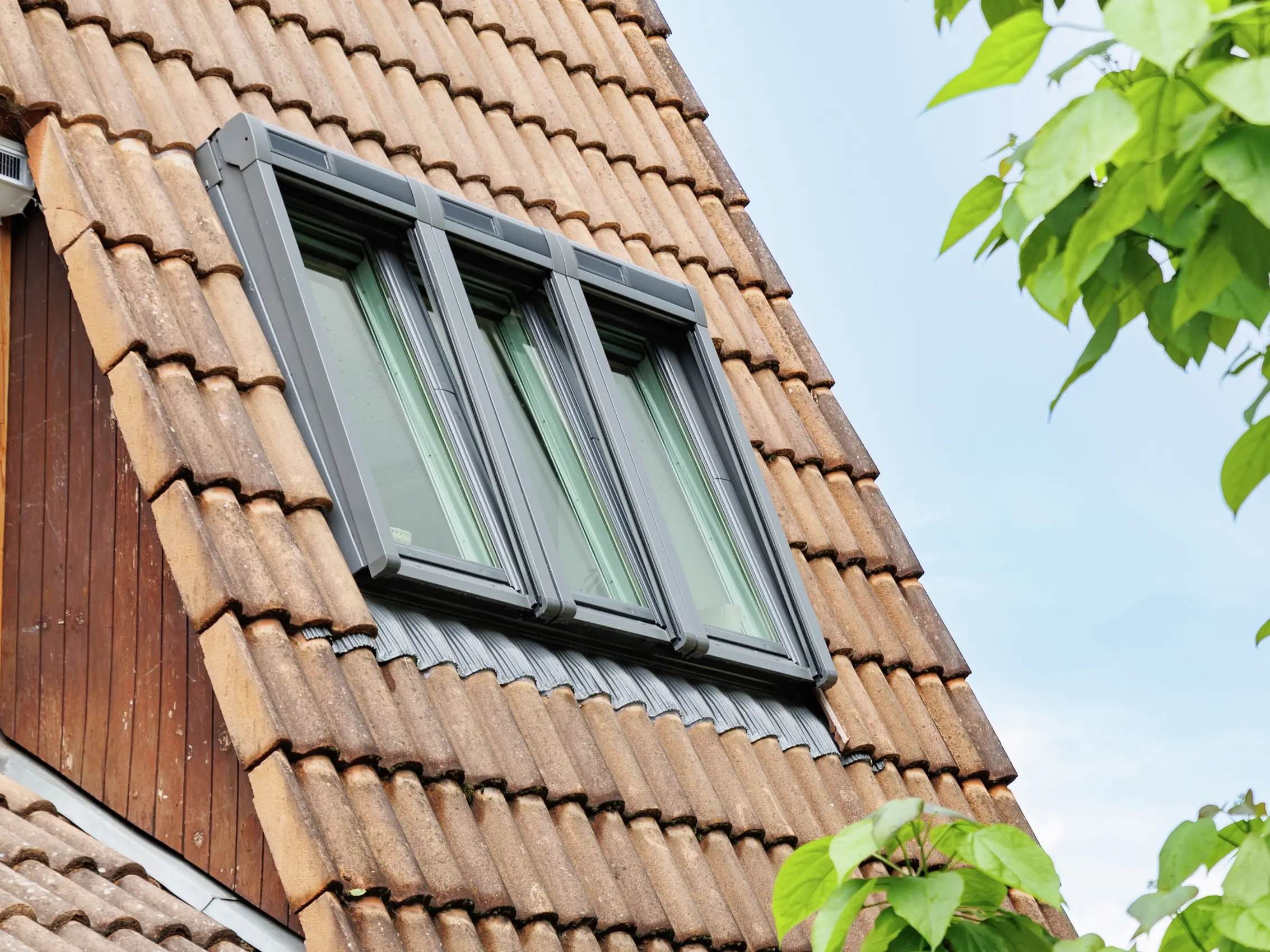 Gros plan d'une fenêtre de toit VELUX sur un toit en tuiles avec des feuilles vertes au premier plan.