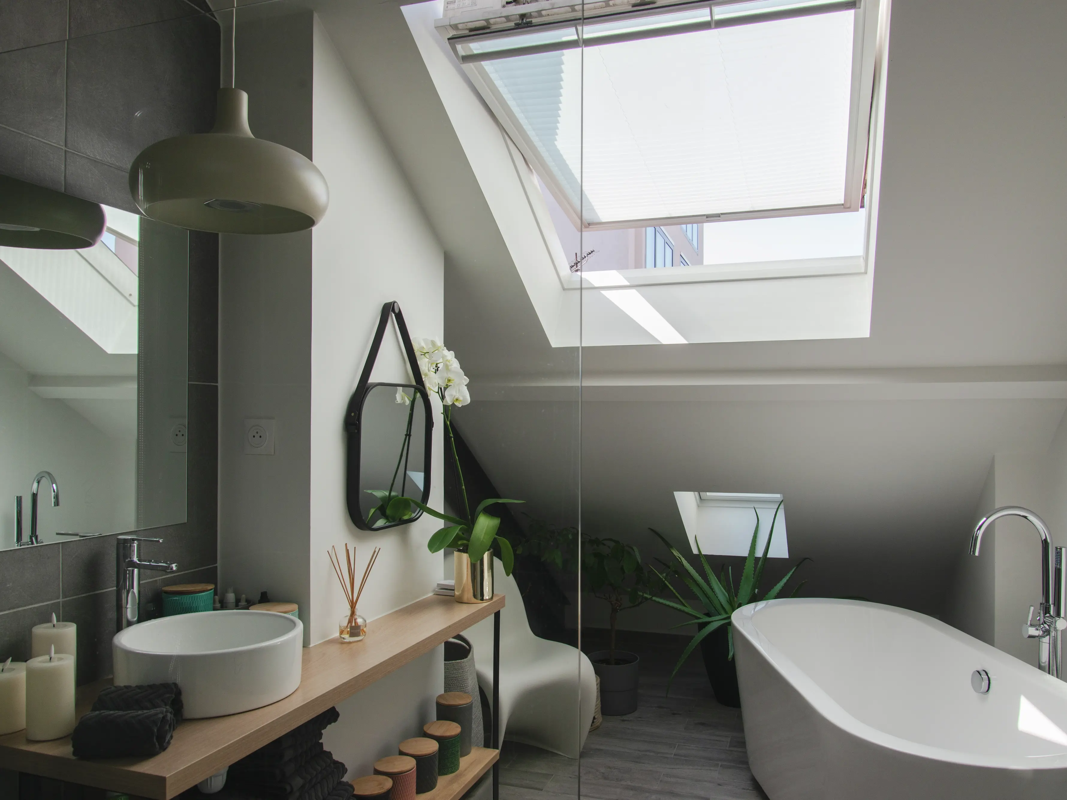 Salle de bain contemporaine avec murs et sol en nuances de gris, une vasque blanche sur un plan de travail en bois, et une fenêtre de toit ouverte laissant entrer la lumière.