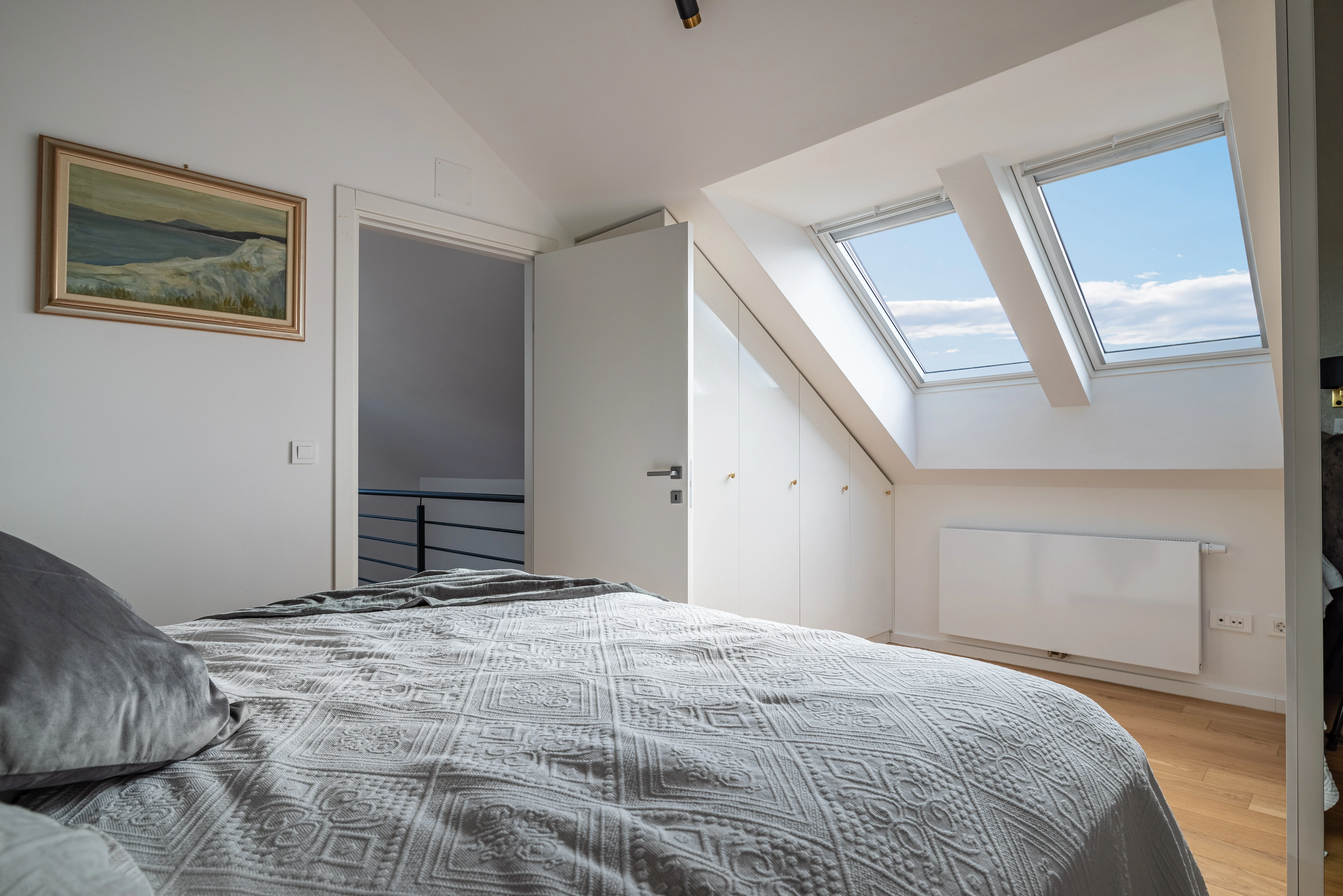 Bēniņu guļamistaba ar slīpiem griestiem un VELUX jumta logiem, pa kuriem ieplūst dabiskā gaisma.