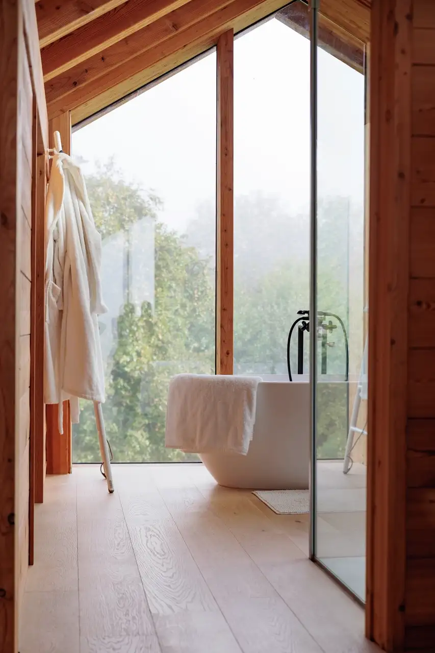 Une salle de bain moderne avec une baignoire blanche autoportante près de grandes fenêtres encadrées de bois offrant une vue paisible sur la verdure, et un peignoir accroché à une échelle en bois.