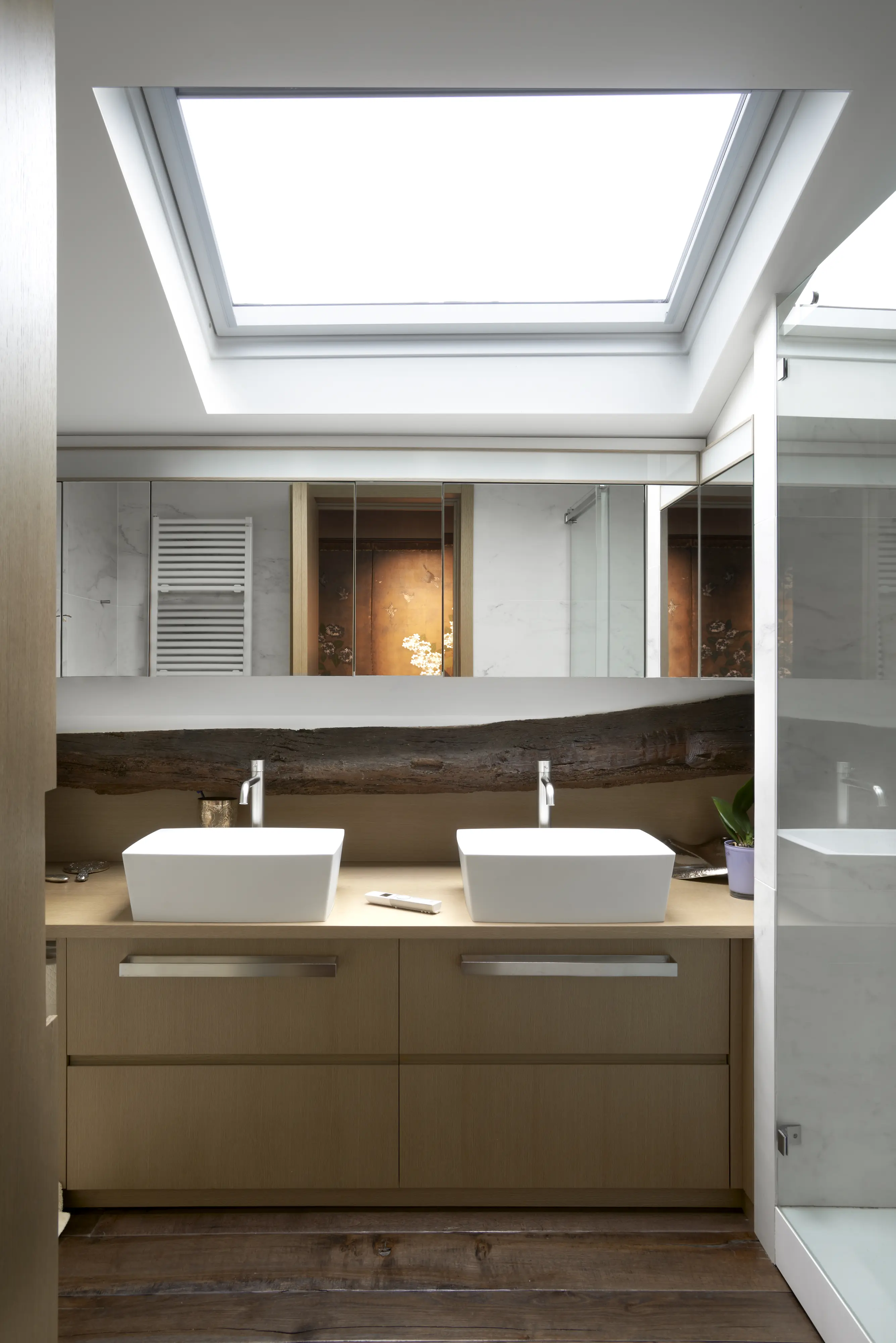 bagno con due lavabi bianchi quadrati da appoggio sopra ad un mobile in legno con maniglie in acciaio satinato, uno specchio lungo tutta la parete, un’ampia doccia con vetro trasparente, una finestra per tetti grande e molto luminosa. 
