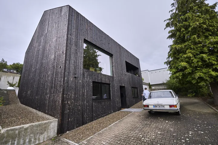 Modernes Einfamilienhaus mit verkohlter Holzfassade und großen Fenstern, klassisches Auto parkt draußen.