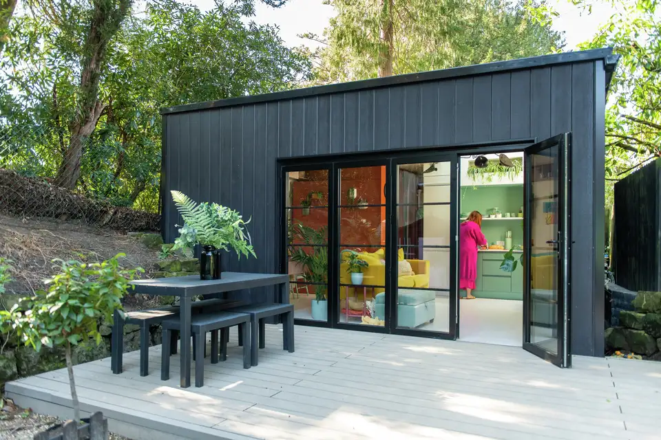 Petit bâtiment moderne noir avec de grandes portes vitrées ouvertes, révélant un intérieur lumineux avec des murs verts. Une personne en rose se tient devant des étagères. À l’extérieur, une terrasse avec une table et des chaises sur des pavés clairs, entourée d’arbres et de végétation.