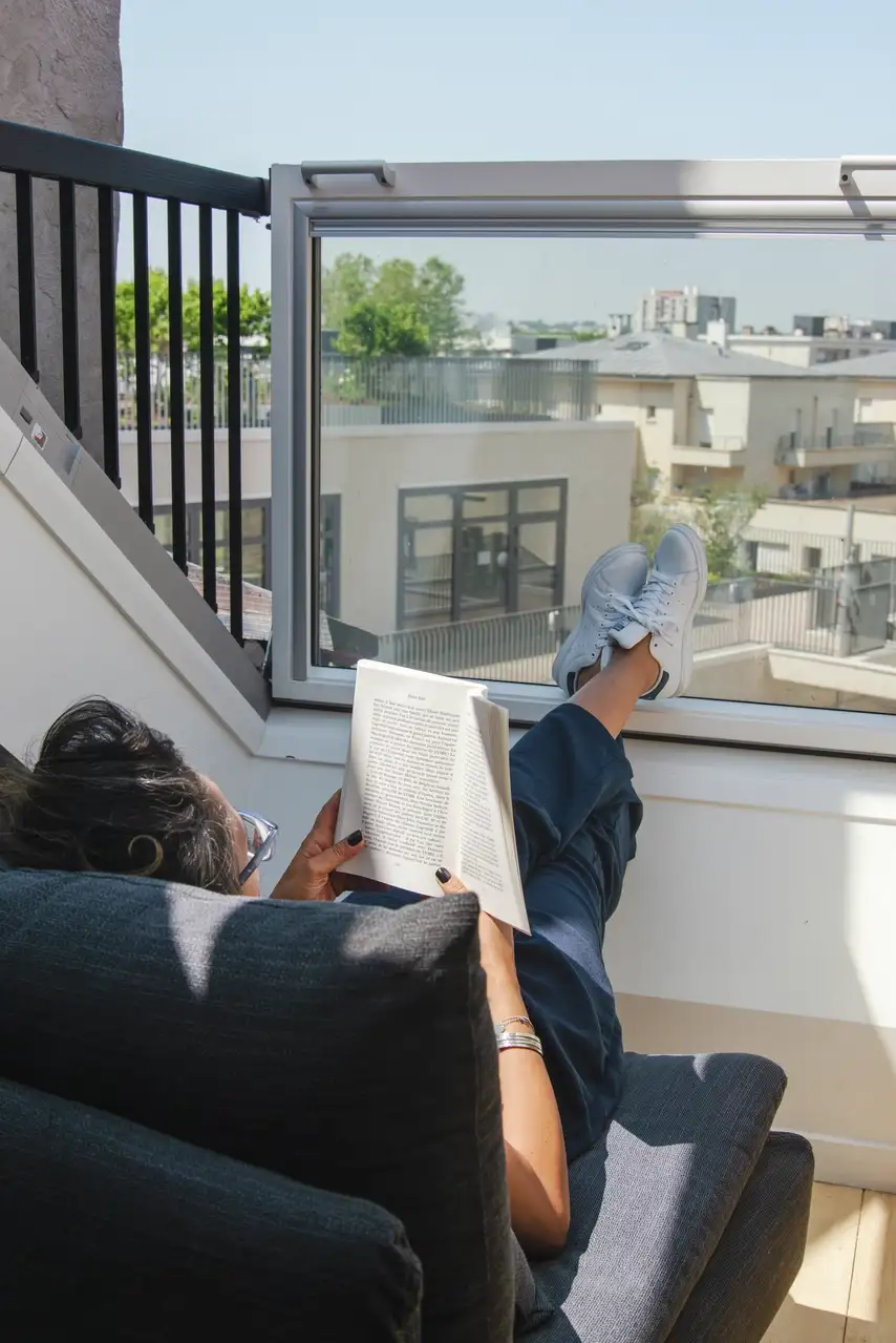 : Une femme se détendant et lisant un livre près d'une fenêtre de toit VELUX ouverte, avec une vue sur la ville et un balcon.