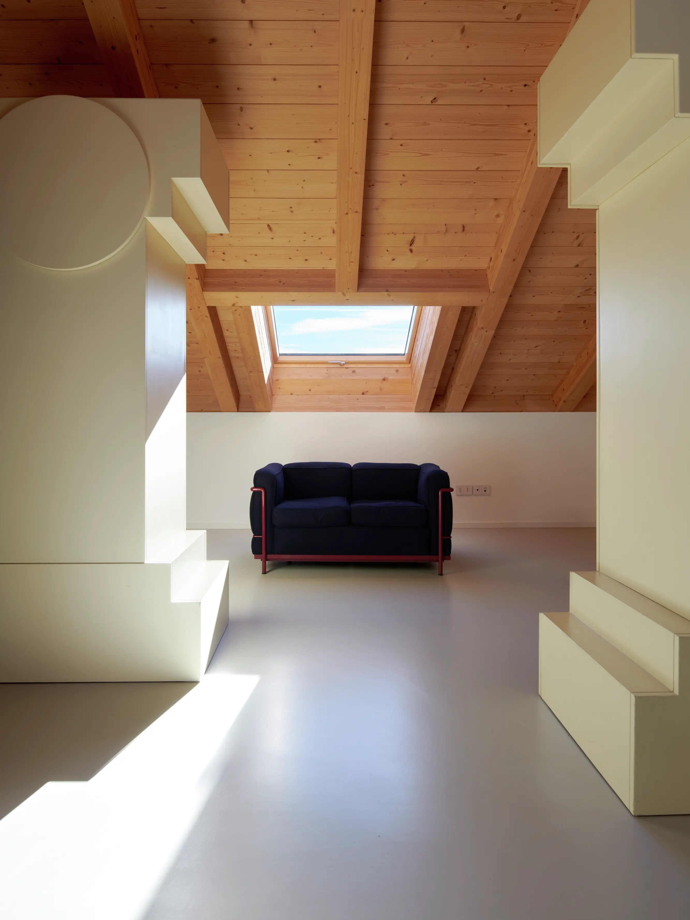 Une pièce lumineuse et aérée avec un plafond en bois, des murs blancs et un canapé noir, éclairée par une fenêtre de toit Velux.