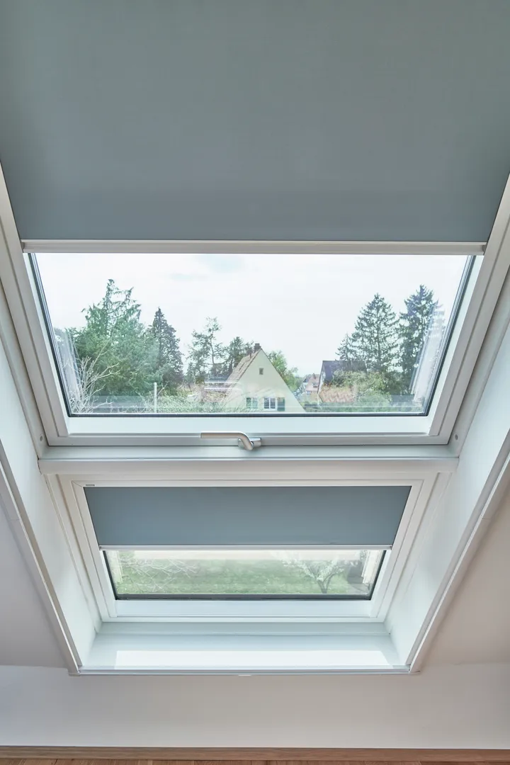 VELUX Dachflächenfenster mit grauer Jalousie, mit Blick auf grüne Landschaft und Haus.