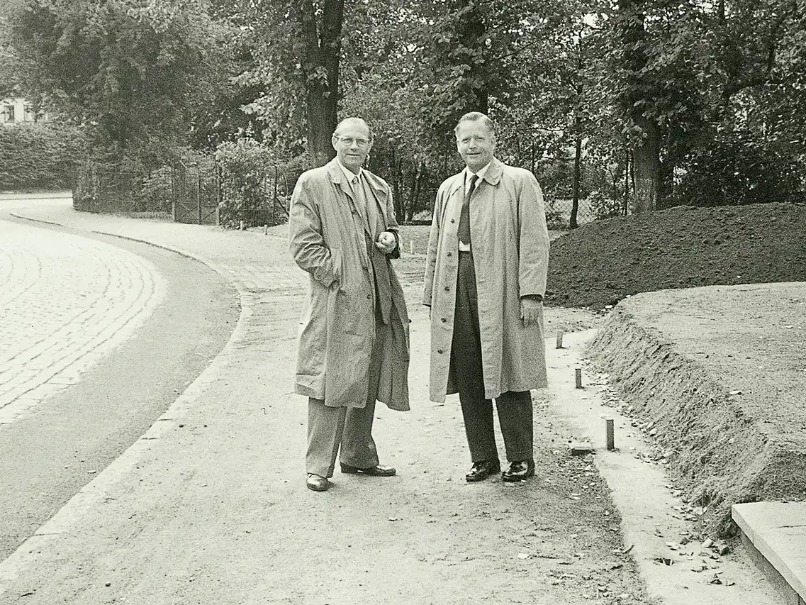 Schwarz-Weiß-Foto von zwei Männern, die auf einem Parkweg sprechen, gekleidet in Vintage-Mänteln.