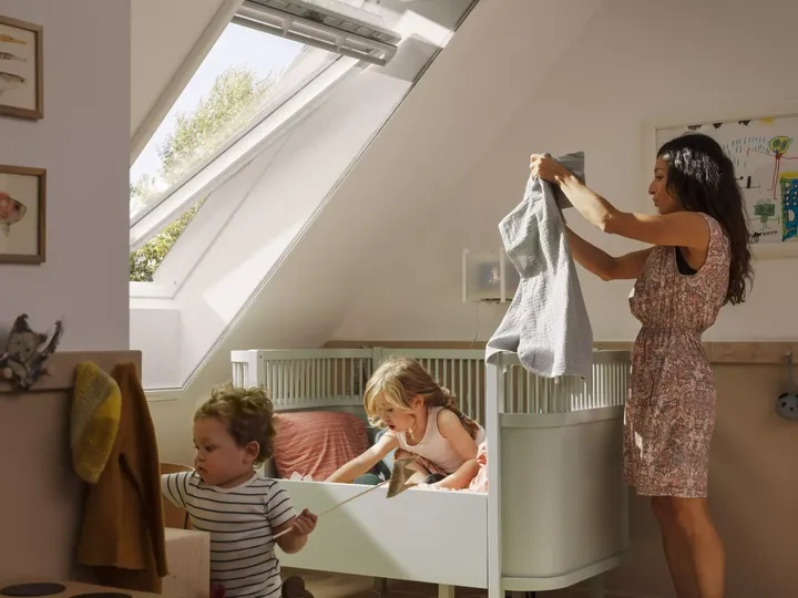 Frau faltet Wäsche während zwei Kleinkinder im Kinderbett unter dem Dachfenster spielen | VELUX Magazin