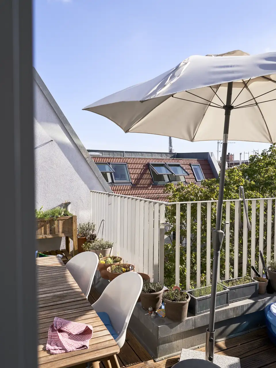Een balkon met houten vloeren en een witte reling. Een open witte parasol biedt schaduw boven een zitgedeelte buiten, dat bestaat uit stoelen met kussens. Op de achtergrond is de bovenkant van een rood bakstenen gebouw met ramen en een schuin dak zichtbaar onder een helderblauwe hemel.