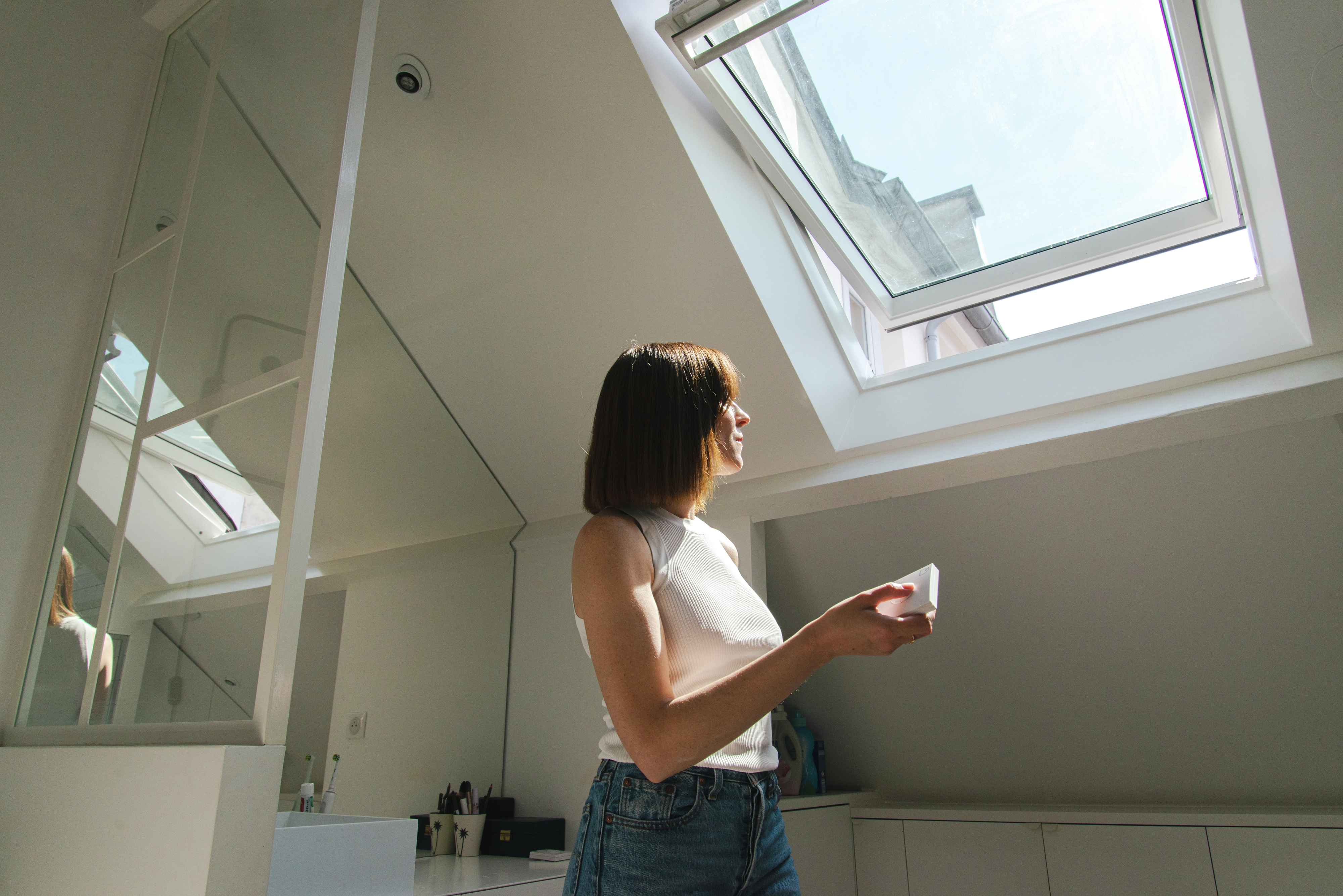 Une femme admire la vue extérieure à travers une fenêtre de toit Velux ouverte, illuminant une pièce mansardée moderne et spacieuse. Cette description met en évidence l’importance de la fenêtre Velux dans le confort intérieur et l’éclairage naturel extérieur.