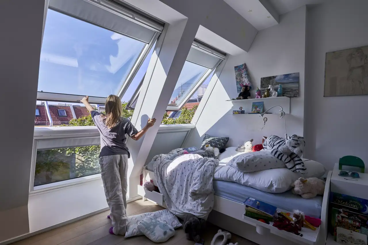 Mädchen öffnet ein VELUX Dachflächenfenster in einem sonnendurchfluteten Dachboden-Schlafzimmer, das mit Spielzeug gefüllt ist.