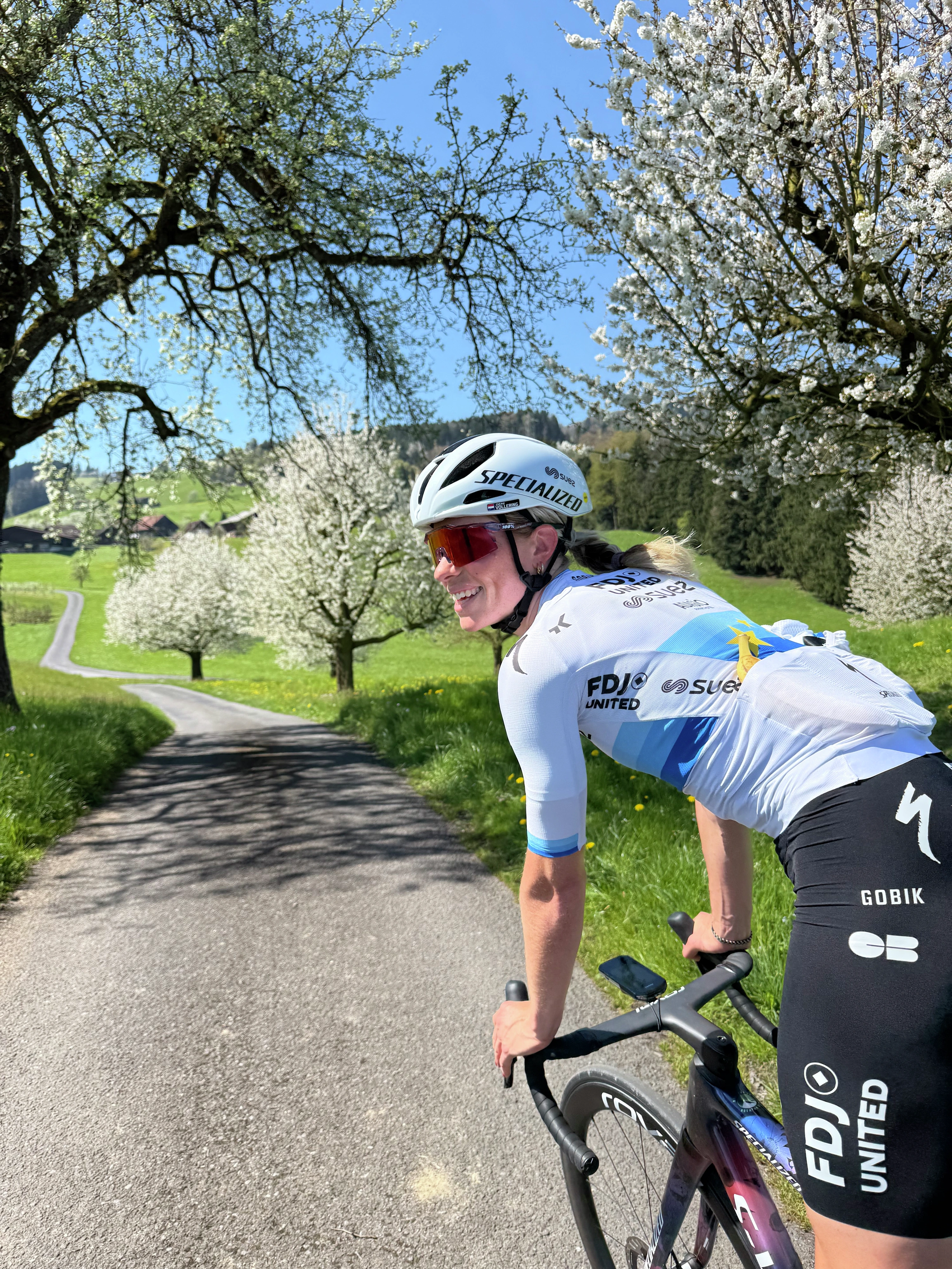 Demi Vollering looks over her shoulder while cycling through a beautiful green landscape with blossoming trees on a sunny day.