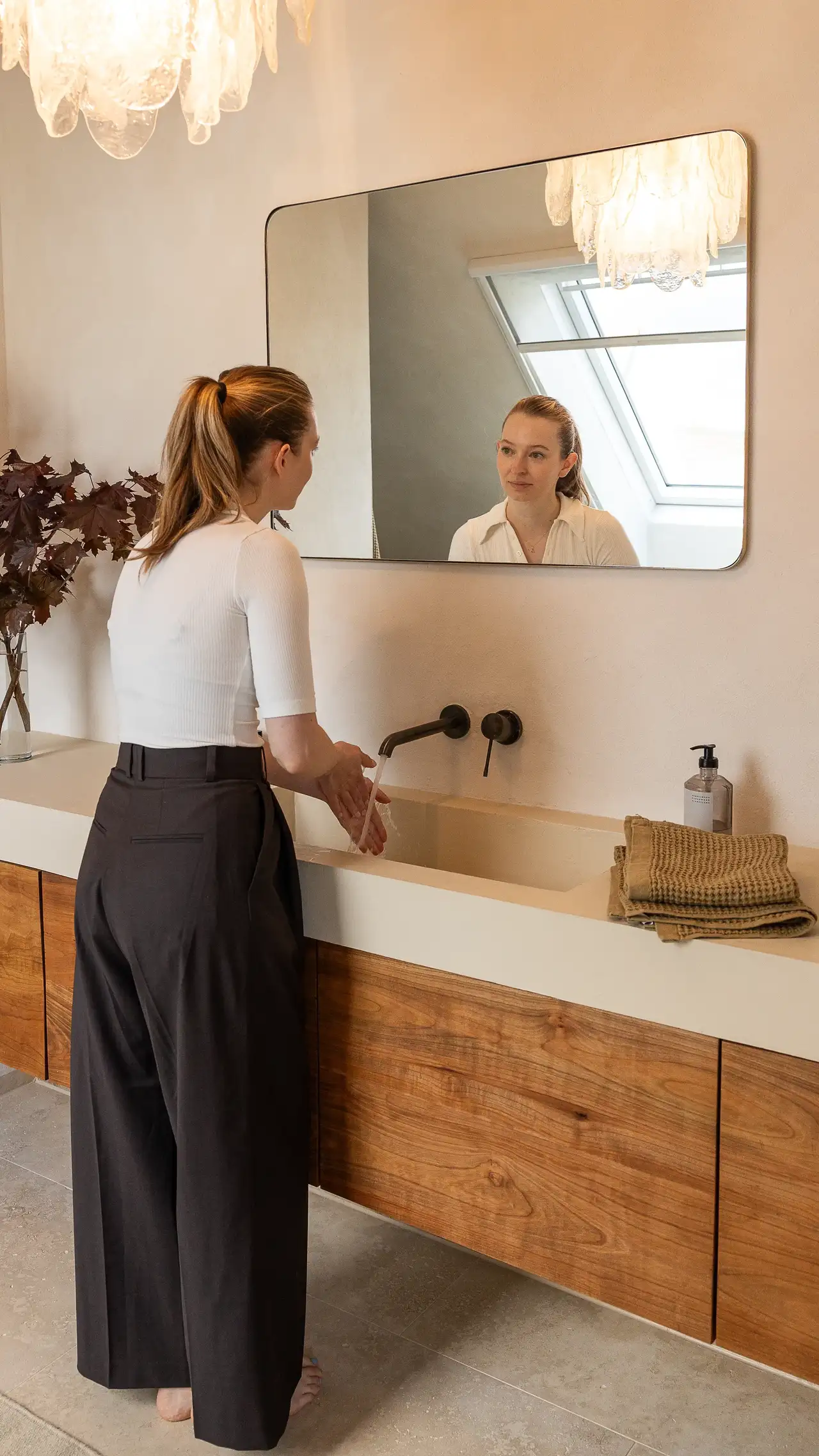Eine Frau wäscht sich die Hände an einem modernen Waschbecken, während sich ein VELUX Dachfenster im Spiegel spiegelt und Tageslicht hereinlässt. | VELUX Magazin