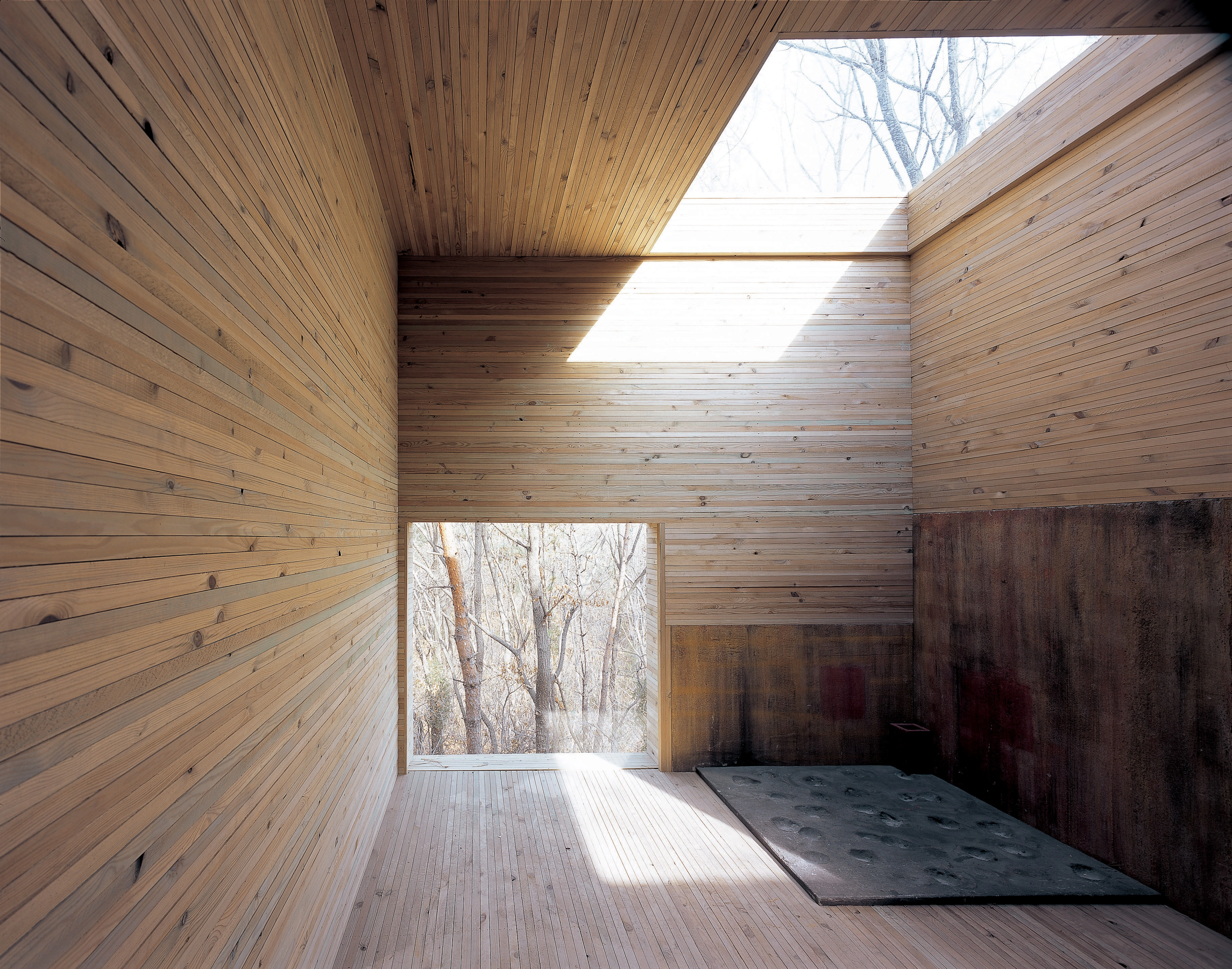 L’immagine mostra uno spazio interno con pareti e pavimento in legno. C’è una grande finestra che permette alla luce naturale di entrare, creando un’atmosfera luminosa. Il design appare minimalista, con linee pulite e nessun mobile visibile, il che potrebbe essere interessante per coloro che apprezzano l’architettura moderna o il design d’interni.