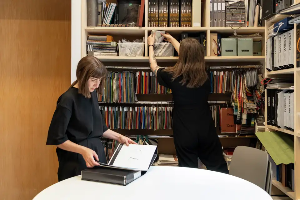 L’immagine mostra due persone in una biblioteca. Una persona è seduta a un tavolo con un libro aperto, apparentemente studiando o leggendo, mentre l’altra persona è in piedi e sta prendendo un libro da uno scaffale. L’ambiente suggerisce un focus su attività accademiche o di ricerca.
